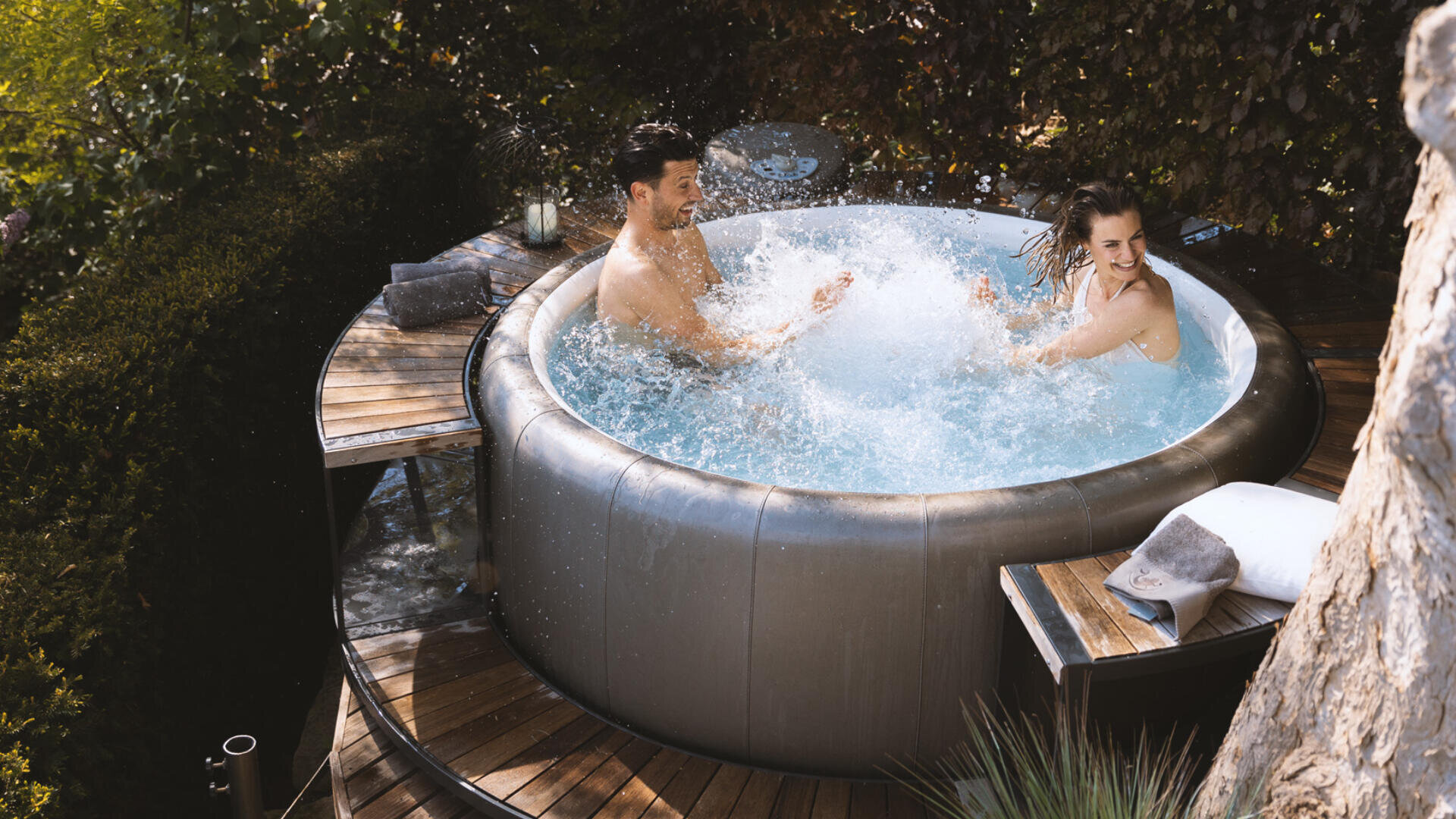 Ein Mann und eine Frau lächeln und bespritzen sich mit Wasser, während sie in einem runden Whirlpool im Freien sitzen, umgeben von Grün und einer Holzterrasse mit Handtüchern und Outdoor-Kissen in der Nähe.