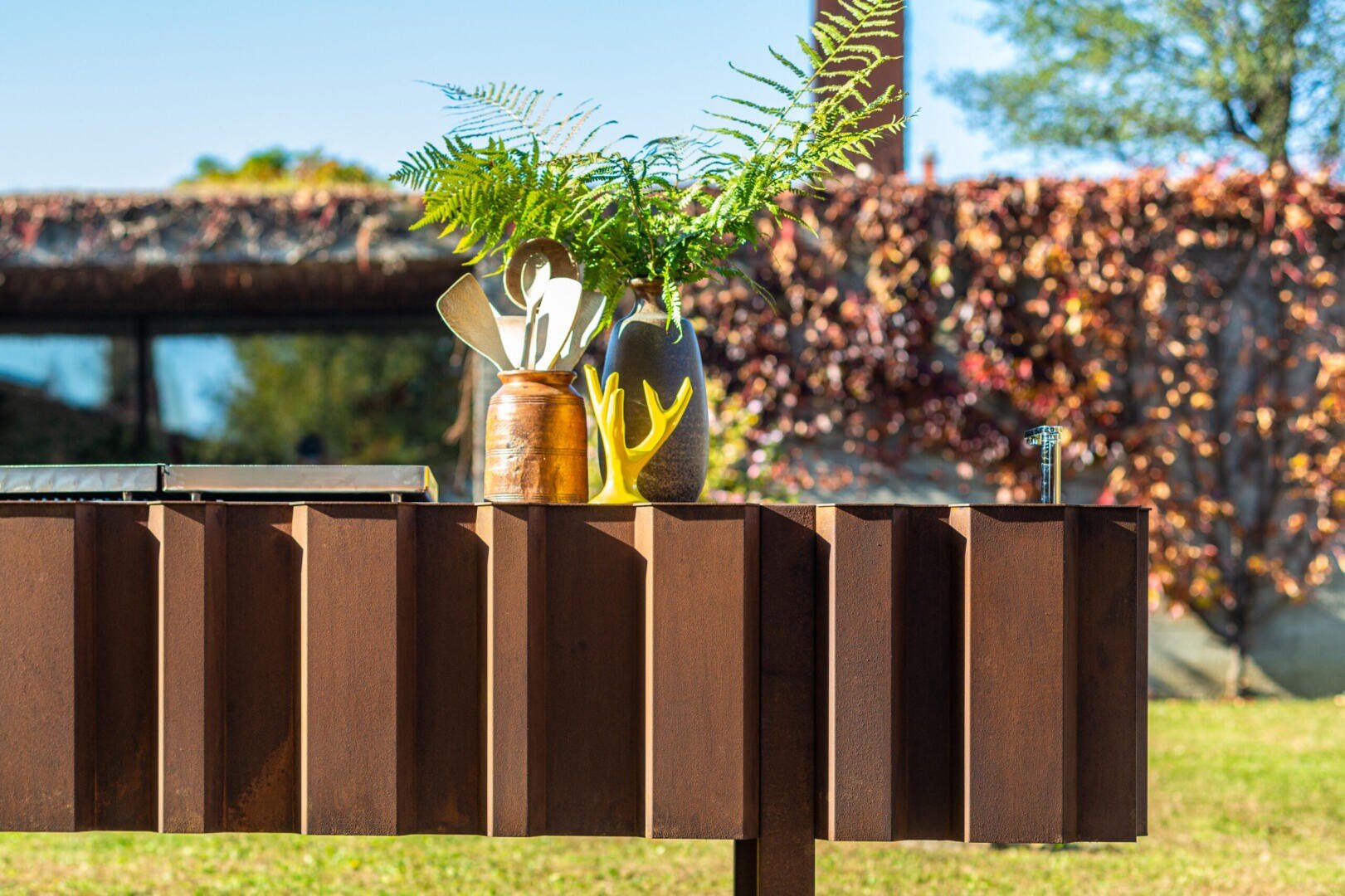 A wooden outdoor table holds a ceramic jar with kitchen utensils and a vase with green ferns. Behind it, there is a wall covered in autumn leaves and trees under a clear sky.