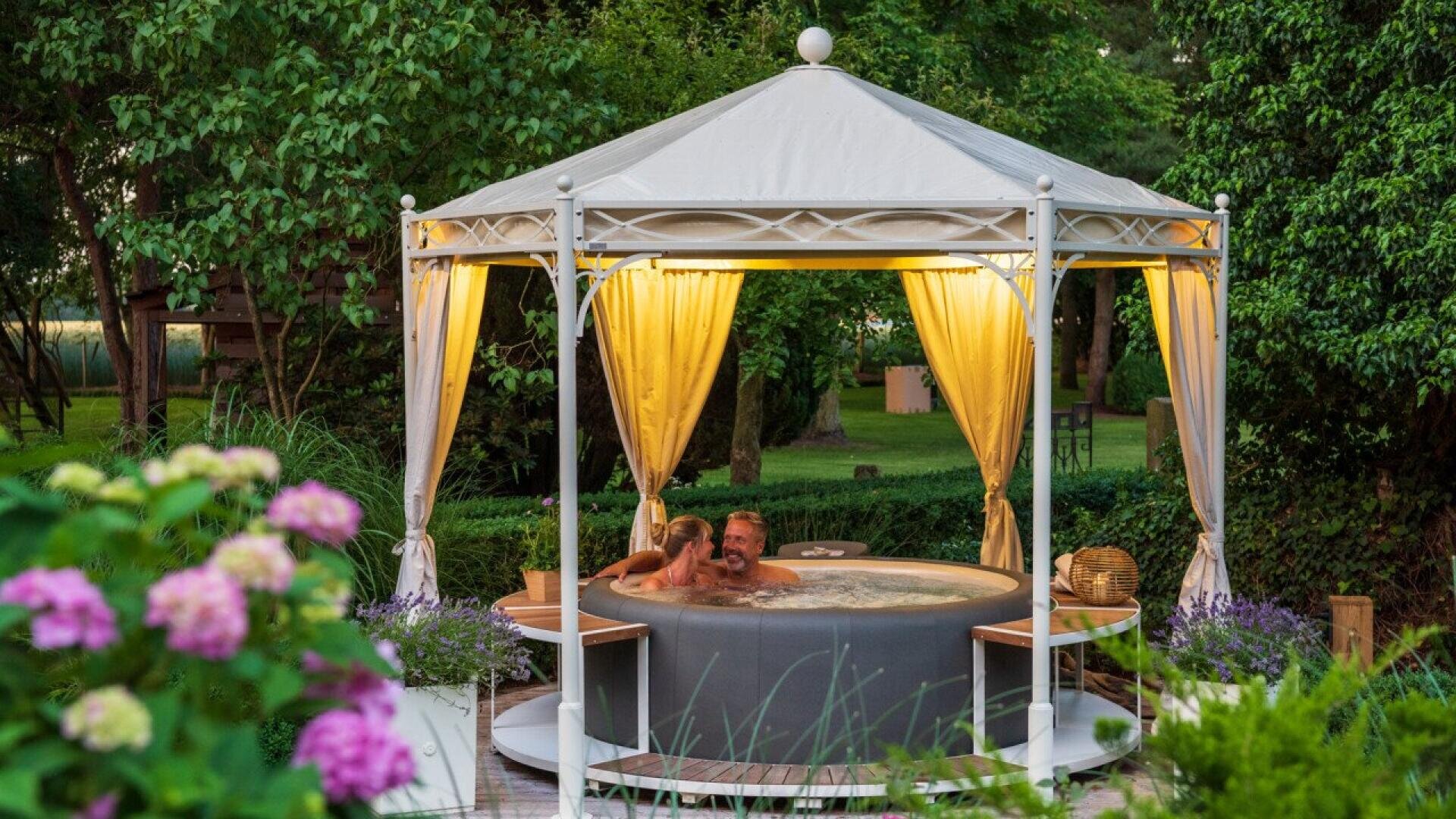 A couple relaxes in a round hot tub under a white gazebo with yellow curtains, surrounded by lush greenery and blooming flowers in a peaceful garden setting.