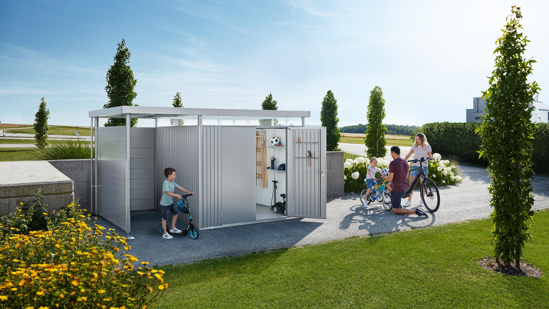 Eine Familie steht draußen vor einem modernen Metallschuppen; ein Junge mit einem Motorroller steht in der Nähe, während ein anderes Kind und ein Erwachsener mit Fahrrädern davor stehen. Die Tür des Schuppens ist offen und zeigt organisierte Sport- und Gartengeräte im Inneren.