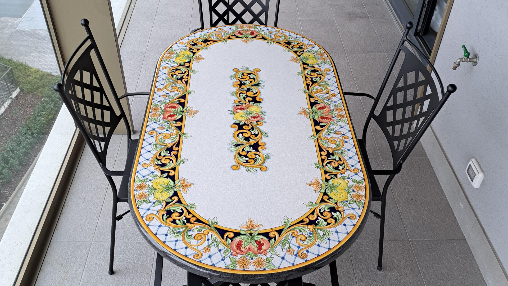 Ein verschnörkelter ovaler Tisch mit buntem Blumen- und geometrischem Muster, umgeben von drei schwarzen Metallstühlen mit vergitterten Rückenlehnen, steht auf einem gefliesten Balkon mit Glasgeländer.