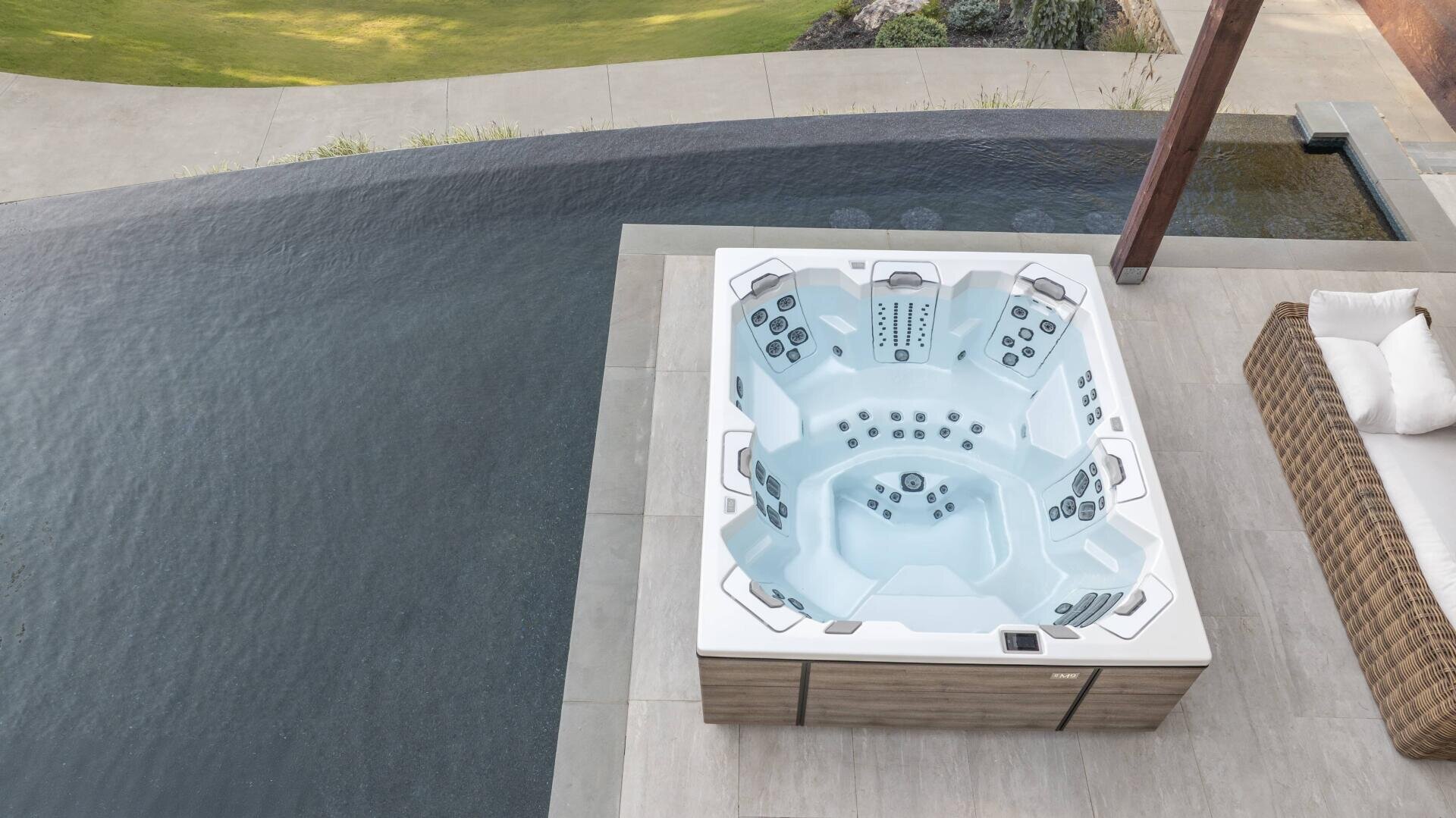 A modern whirlpool with several seats is located next to an infinity pool on a tiled terrace, next to a wicker sofa with white cushions and a view of the green grass beyond.