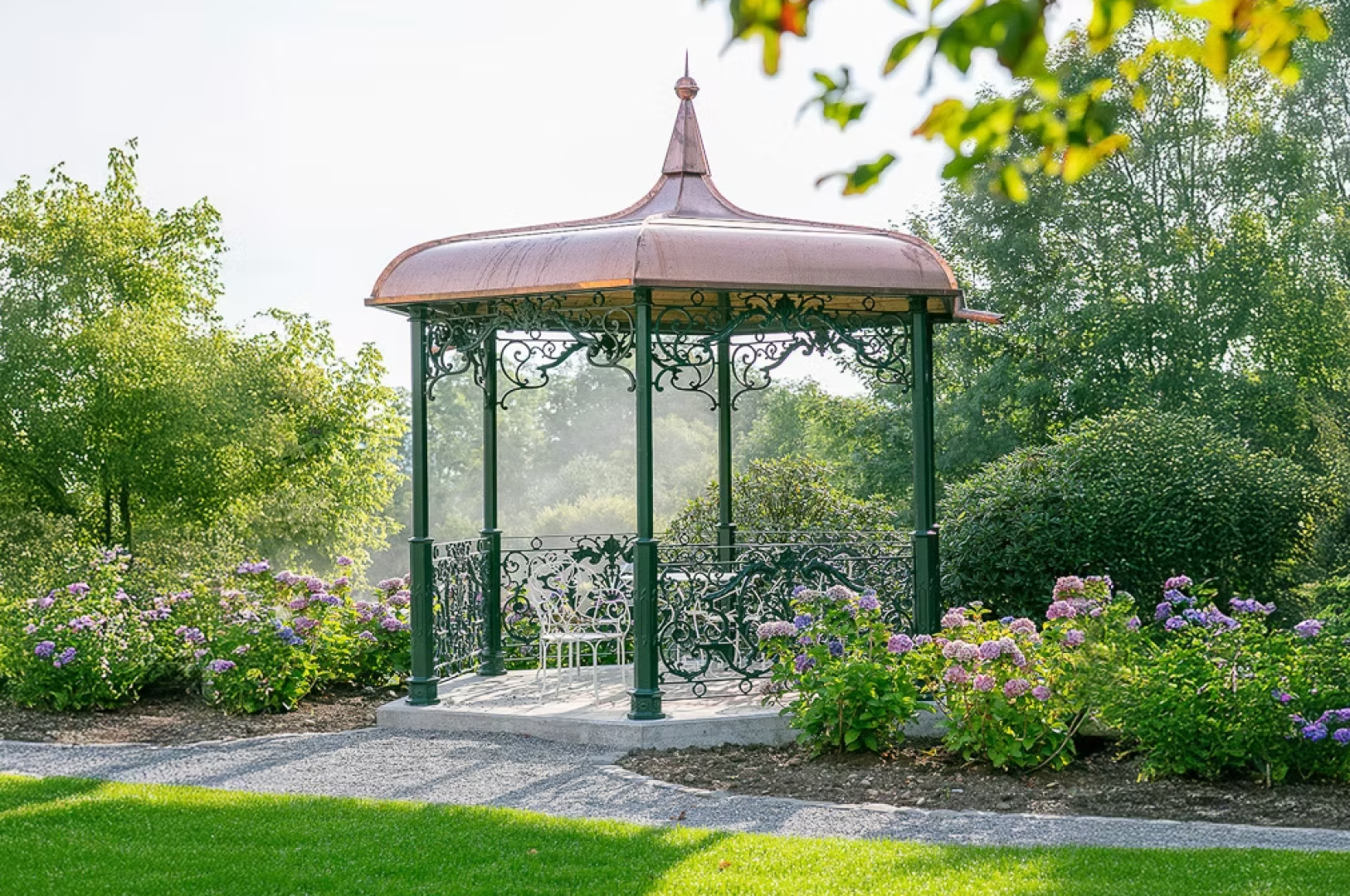 Ein dekorativer Metallpavillon mit Kupferdach steht in einem Garten, umgeben von blühenden lila Blumen und grünen Sträuchern, mit einer weißen Bank im Inneren und Bäumen im Hintergrund.
