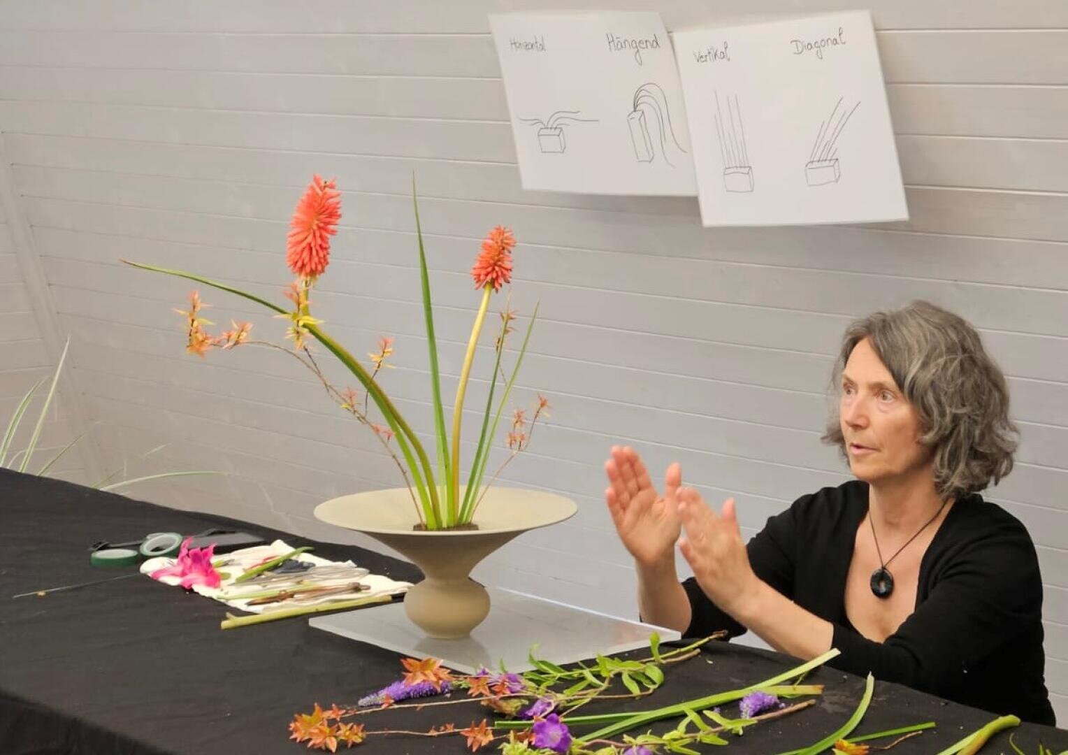 Eine Frau demonstriert das Arrangieren von Blumen an einem Tisch, der mit Werkzeugen und Blumen bedeckt ist. Hinter ihr sind an der Wand Diagramme mit beschrifteten Skizzen von Blumenbindetechniken zu sehen.