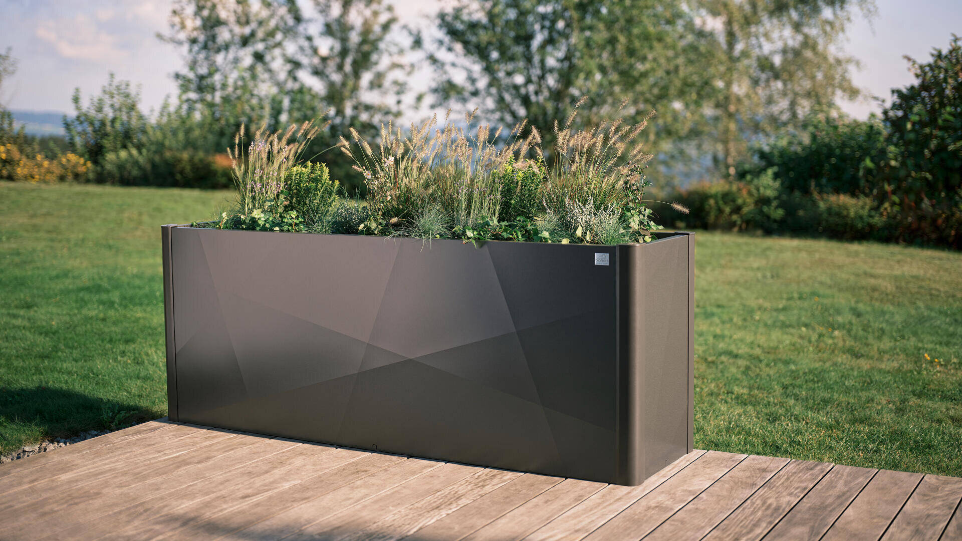 Ein moderner, rechteckiger Metall-Pflanzkasten, gefüllt mit Gräsern und Pflanzen, steht an einem sonnigen Tag auf einer Holzterrasse in einem grasbewachsenen Garten mit Bäumen im Hintergrund.