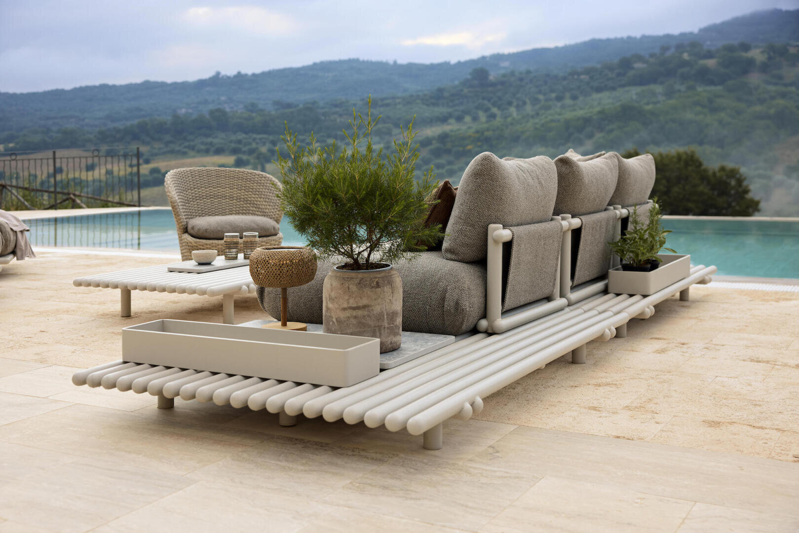 Ein moderner Lounge-Bereich im Freien mit gepolsterten Sitzgelegenheiten, Topfpflanzen und einem Beistelltisch aus Weidengeflecht befindet sich auf einer Steinterrasse neben einem Infinity-Pool mit Blick auf grüne Hügel und einen wolkenverhangenen Himmel.