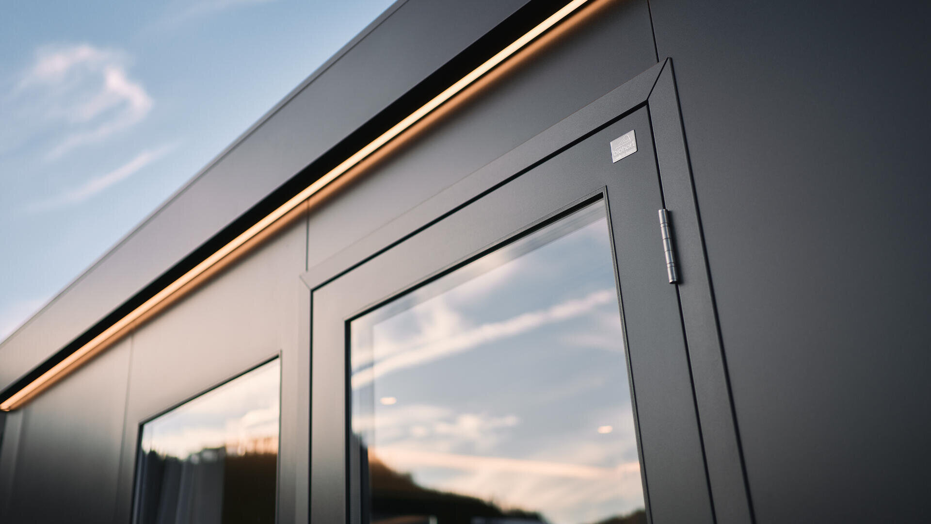 Nahaufnahme einer modernen dunkelgrauen Tür und eines Fensters mit spiegelndem Glas, eingebettet in eine schlichte Gebäudehülle. Ein durchgehendes LED-Lichtband verläuft über der Tür unter einem blauen Himmel mit Wolkenfetzen.