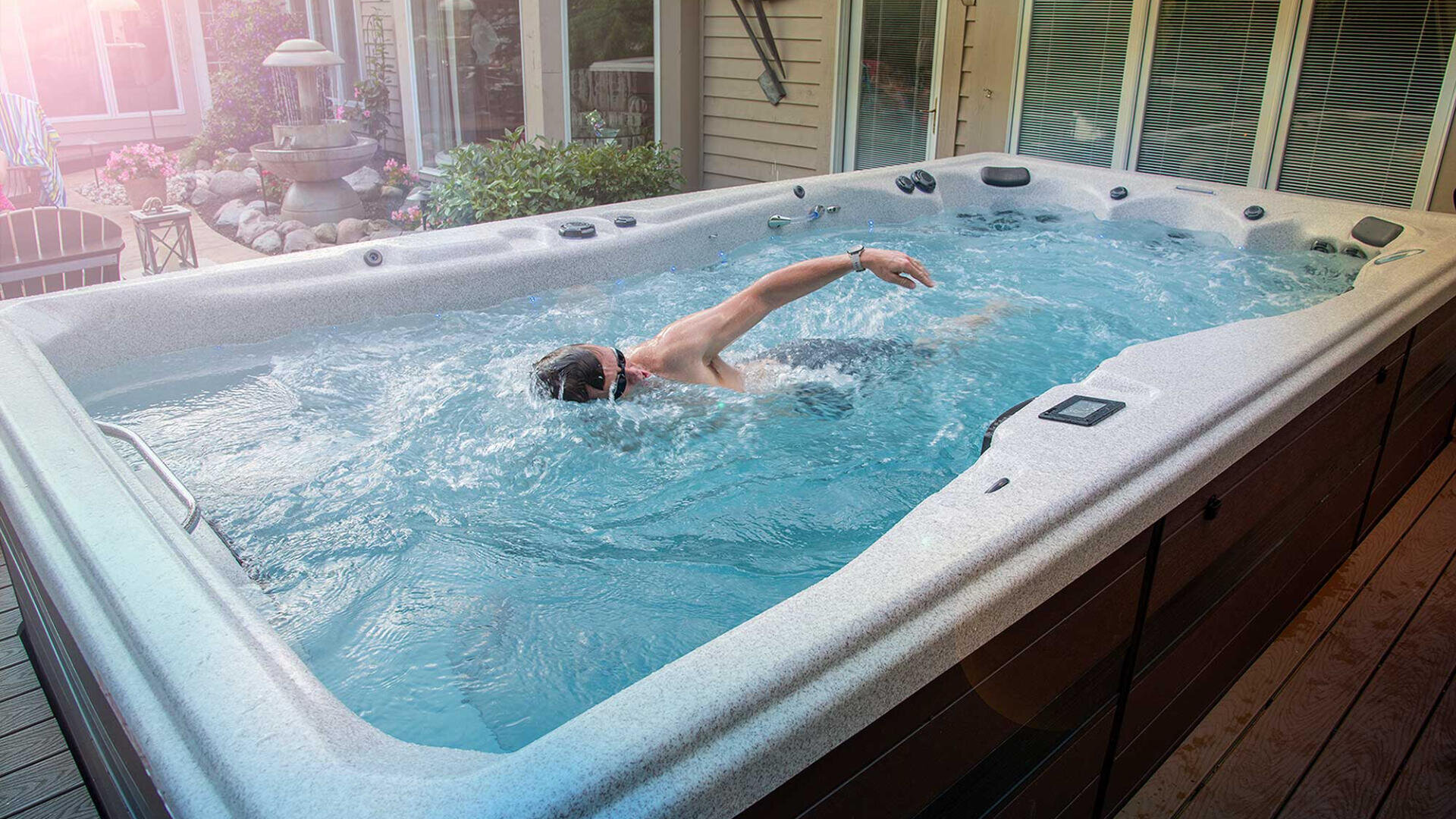 Eine Person schwimmt in einem großen Whirlpool im Freien mit einer Schwimmbrille im vorderen Bereich. Der Whirlpool befindet sich auf einer Terrasse neben einem Haus mit Glastüren und umliegenden Pflanzen.