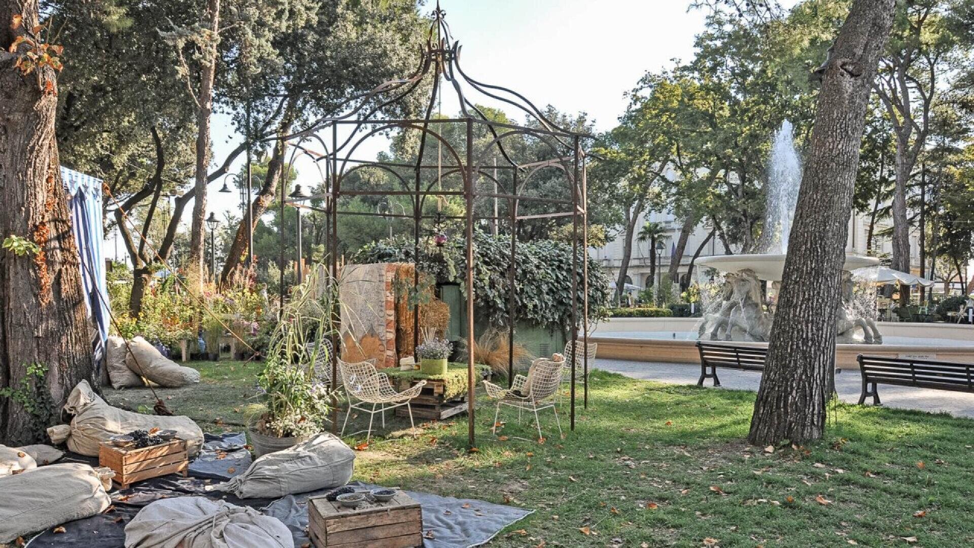 A garden scene with a metal gazebo frame, two chairs, potted plants, bean bags, wooden crates, and a fountain in the background, surrounded by trees and greenery on a sunny day.