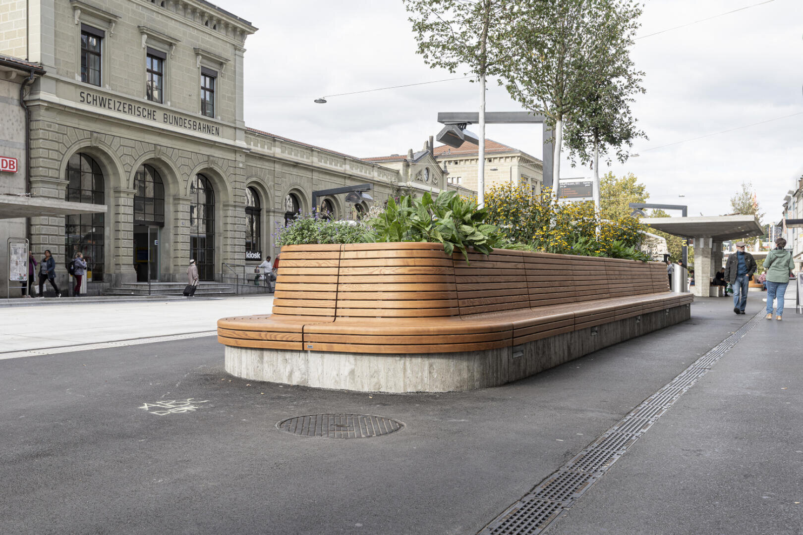 Eine lange, geschwungene Holzbank mit eingebauten Pflanzgefäßen und Grünpflanzen steht auf einem Gehweg in der Nähe des Bahnhofs der Schweizerischen Bundesbahnen. In der Nähe gehen Menschen spazieren und im Hintergrund ist die Steinfassade des Bahnhofs zu sehen.