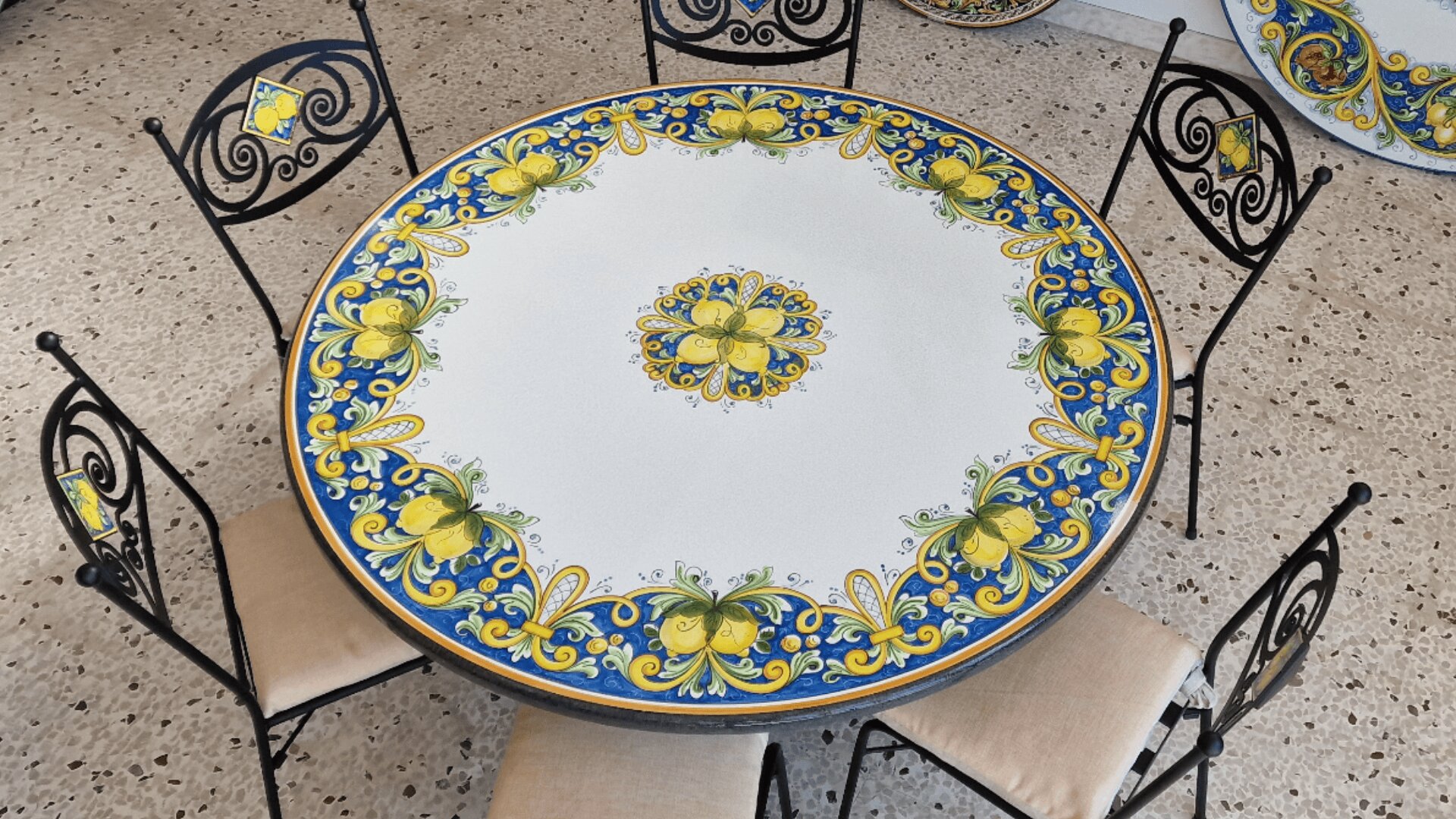 A round table with a colorful ceramic top decorated with yellow lemons and blue floral patterns, surrounded by five black wrought iron chairs with beige cushions and matching lemon designs on the backrests.