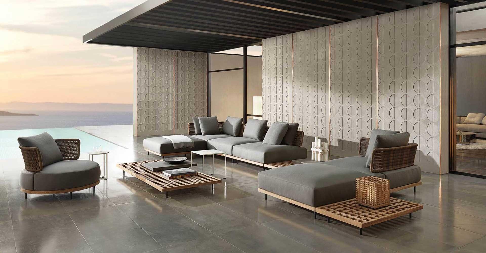 Modern outdoor lounge area with gray upholstered sofas, wooden coffee tables and a round chair by the pool with sea views and geometric patterned wall panels in the background.