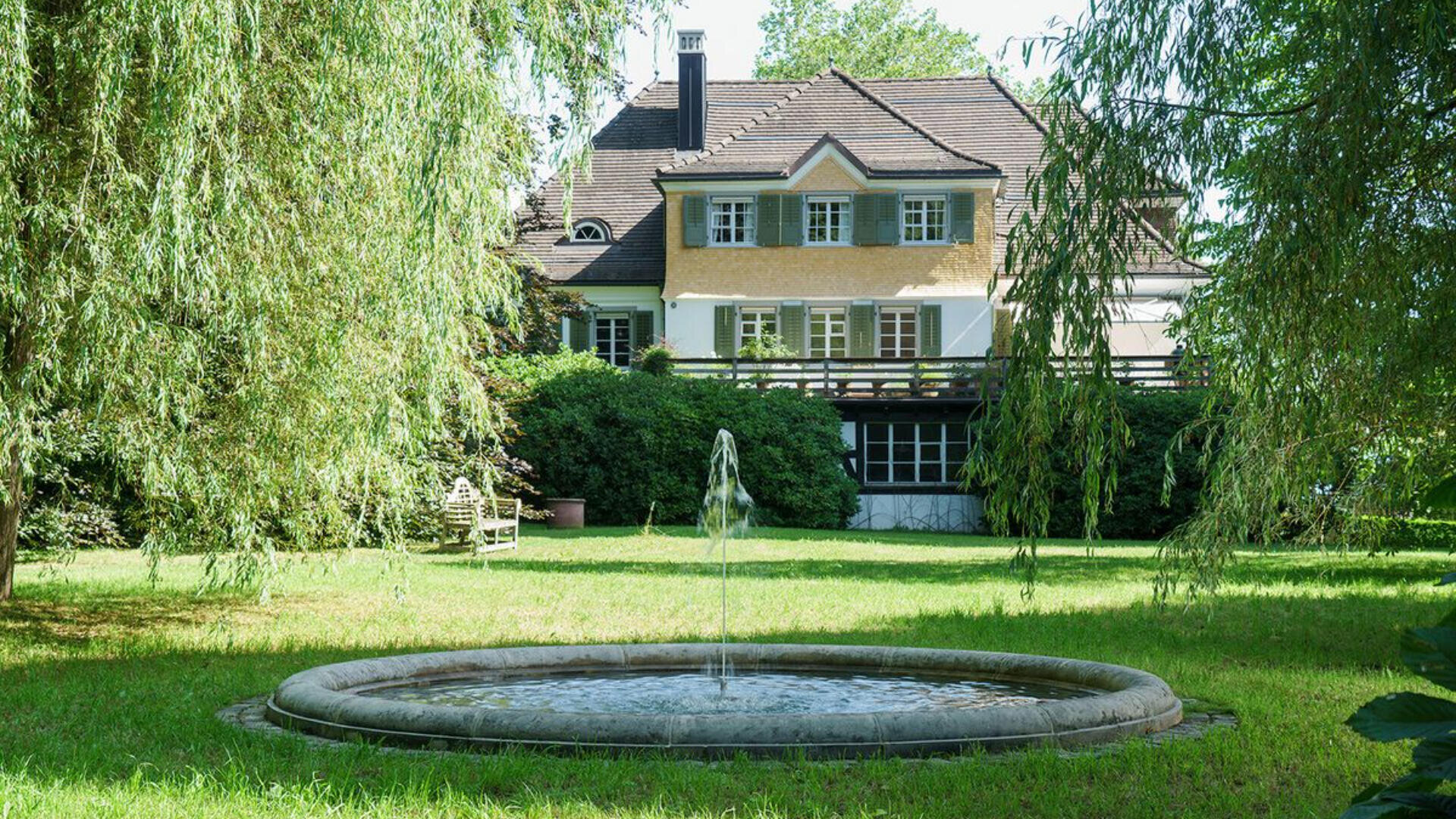 Ein runder Steinbrunnen steht auf einer von Bäumen umgebenen grünen Wiese. Im Hintergrund steht ein großes zweistöckiges Haus mit grünen Fensterläden und einem schrägen Dach. Das Sonnenlicht fällt durch die Blätter.