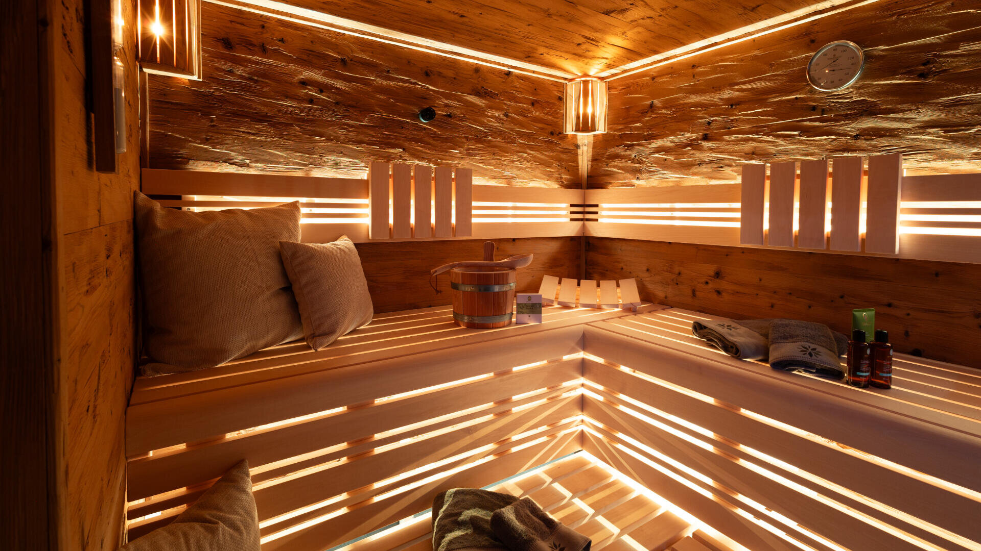Eine gemütliche Holzsauna mit sanfter Beleuchtung, mit Kissen ausgelegten Bänken, einem Eimer und einer Kelle, einem Handtuch, Flaschen und einer Zeitschrift auf der Bank. An der Wand darüber ist eine Uhr angebracht. Die Atmosphäre ist warm und einladend.