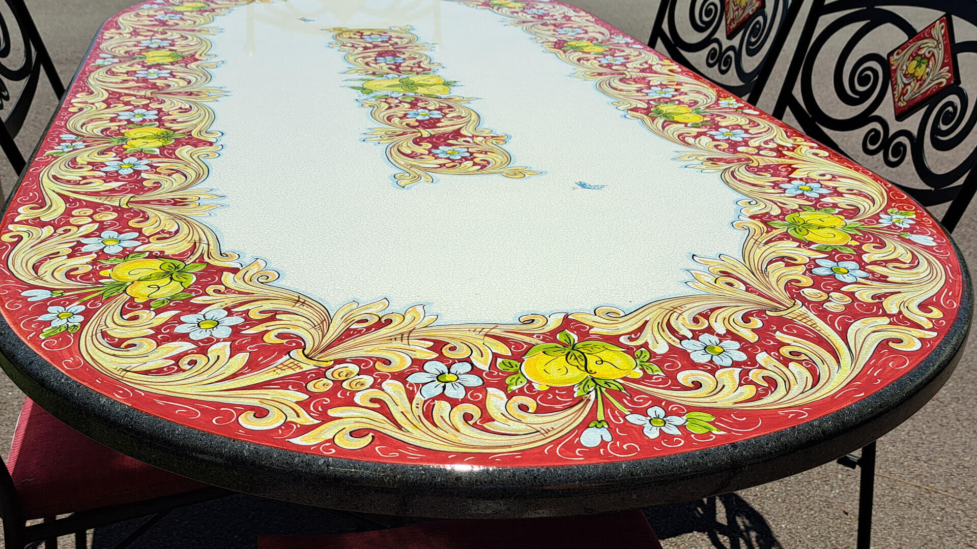 Ein ovaler Tisch mit einem dekorativen roten, gelben und cremefarbenen Blumenmuster, umgeben von verzierten schwarzen Stühlen mit roten und gelben Details. Der Tisch steht im Freien, und das Sonnenlicht hebt die glänzende Oberfläche hervor.