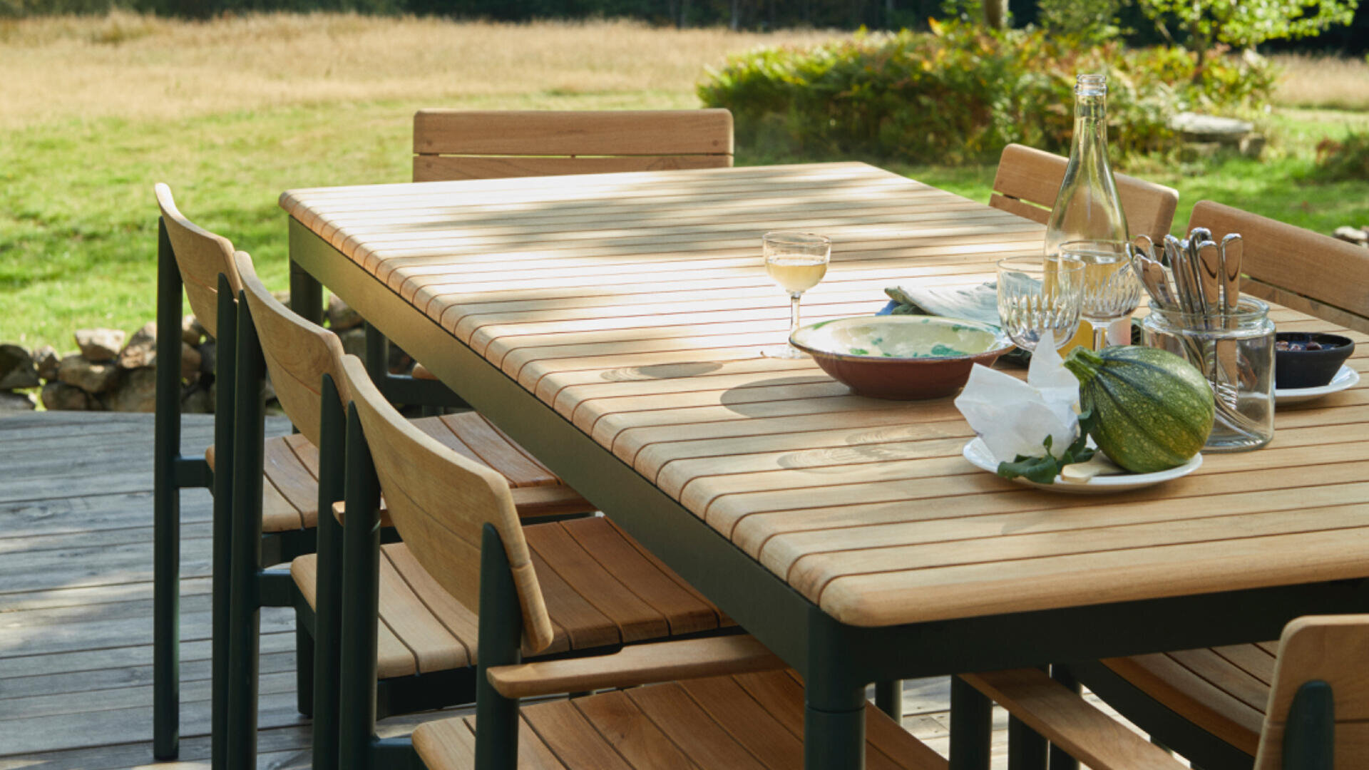 Ein hölzerner Esstisch im Freien mit passenden Stühlen auf einer Terrasse, mit einem Glas Weißwein, Glasflaschen, Geschirr, Besteck und einem Kürbis, mit Gras und Bäumen im Hintergrund.