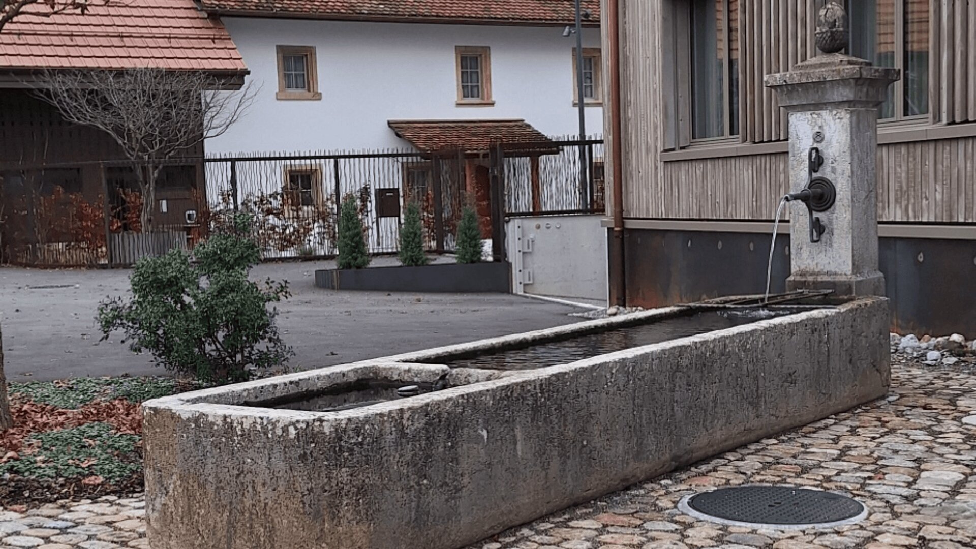 Ein rechteckiger steinerner Wassertrog mit einer vertikalen Fontäne steht auf einer Kopfsteinpflasterfläche neben einem modernen Holzgebäude. Das Wasser fließt aus einem Auslauf in den Trog. Im Hintergrund sind weitere Gebäude und ein eingezäunter Hof zu sehen.