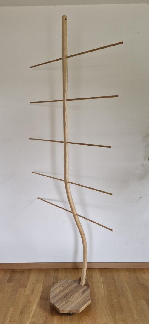 Ein hölzerner Garderobenständer mit einem gebogenen Mittelpfosten, sechs horizontalen Stangen, die sich abwechselnd auf beiden Seiten erstrecken, und einem achteckigen Sockel, der auf einem Holzboden vor einer schlichten weißen Wand steht.