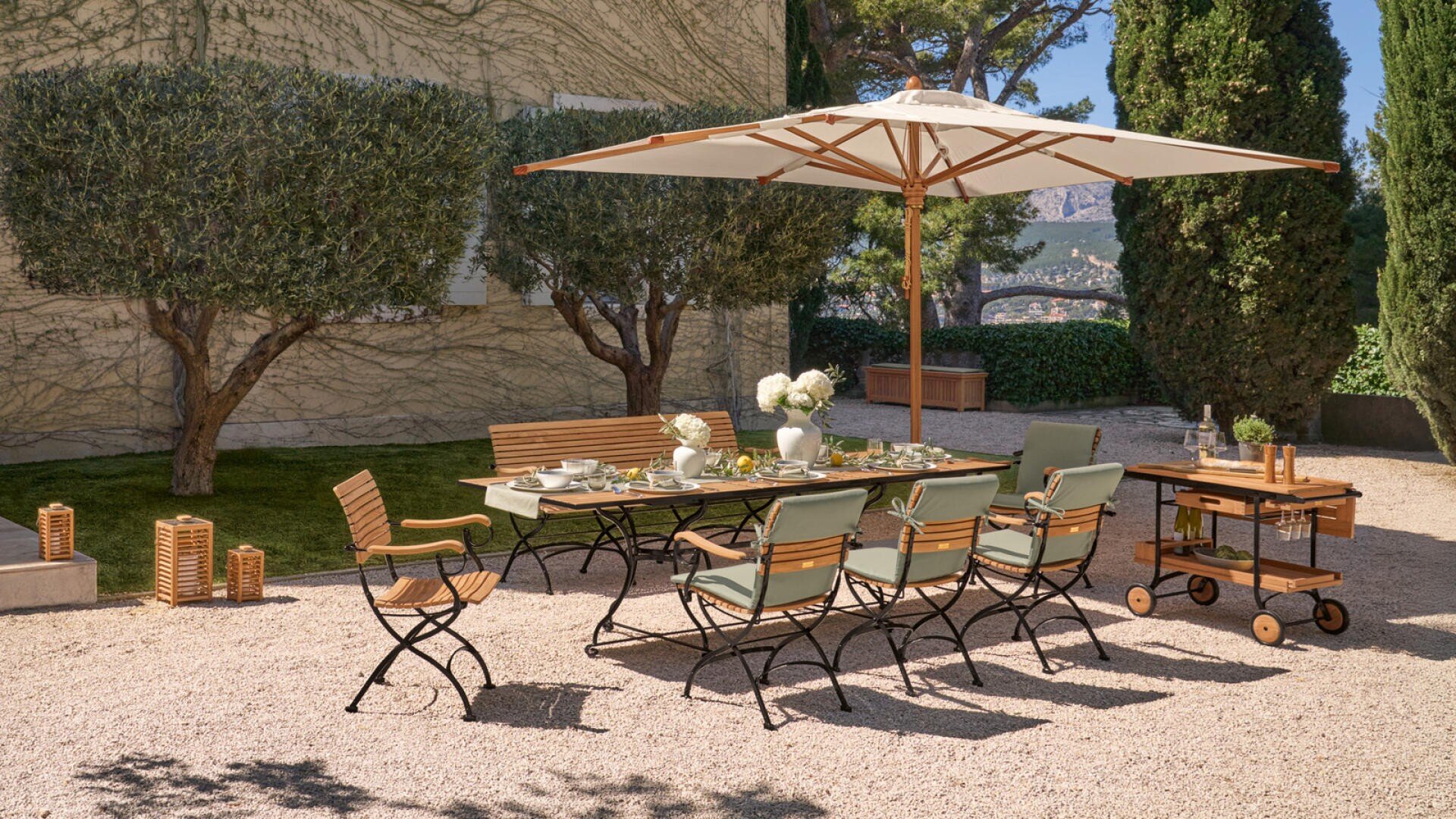 Essbereich auf der Terrasse mit einem Tisch aus Holz und Metall, sechs Stühlen und einer Bank unter einem großen beigen Sonnenschirm. Der Tisch ist für eine Mahlzeit gedeckt, umgeben von Grün und Bäumen auf einer Kiesfläche.