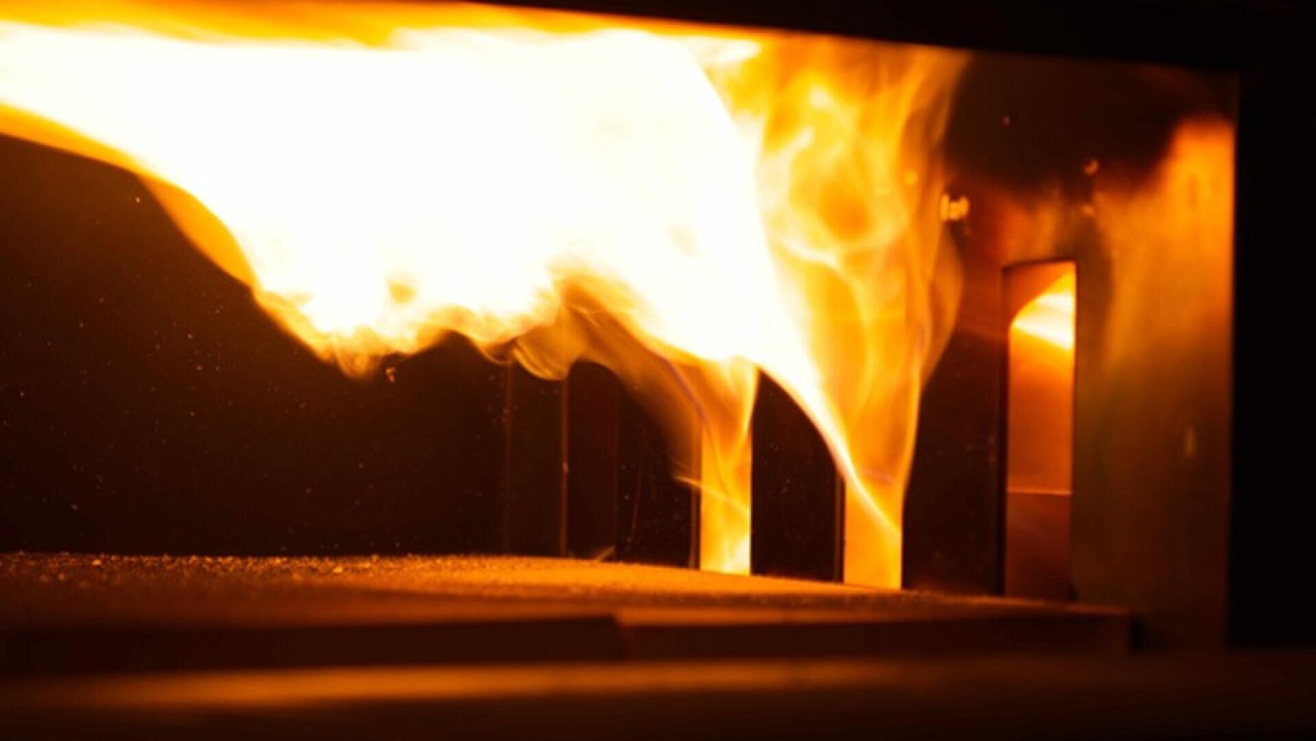 A bright orange and yellow flame burns intensely inside a furnace or industrial oven, casting light and shadows on the dark metal interior.
