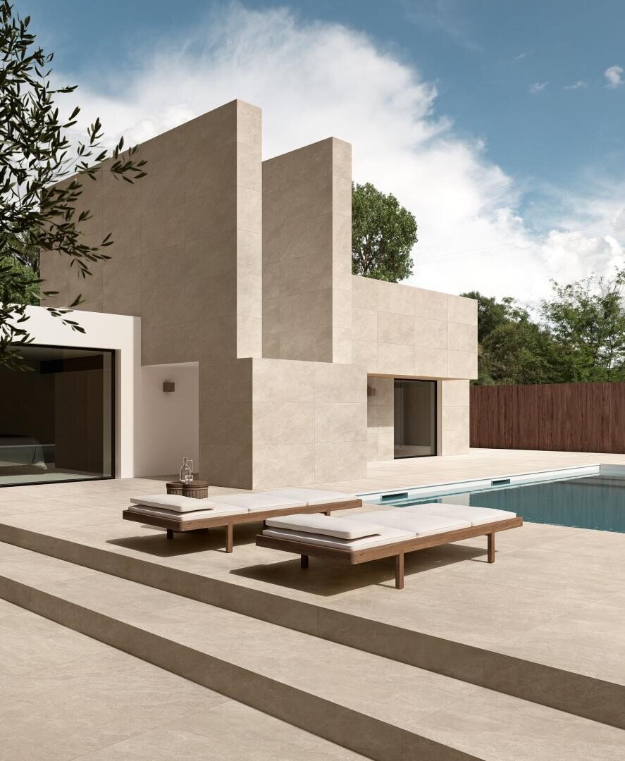 Modernes, minimalistisches Haus mit beigefarbener Steinfassade, großen geometrischen Formen und hohen Fenstern; zwei gepolsterte Holzsessel stehen neben einem rechteckigen Swimmingpool auf einer geräumigen Terrasse unter einem teilweise bewölkten Himmel.