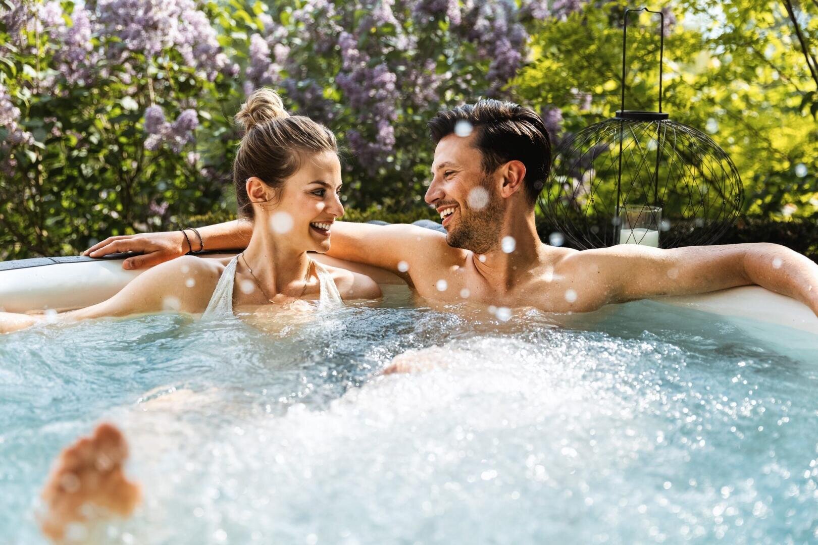 Ein lächelnder Mann und eine lächelnde Frau entspannen sich gemeinsam in einem Whirlpool im Freien, umgeben von Grün und Fliederblüten, und genießen einen sonnigen Tag. Blasen steigen im Wasser zwischen ihnen auf, während sie sich gegenseitig anschauen.