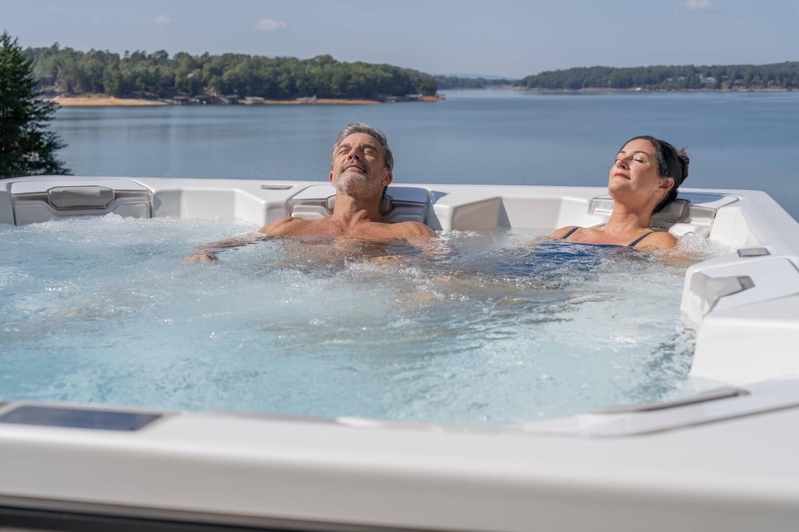 A man and a woman relax in a hot tub overlooking a picturesque lake with trees in the background under a clear sky. Both have their eyes closed and appear peaceful.