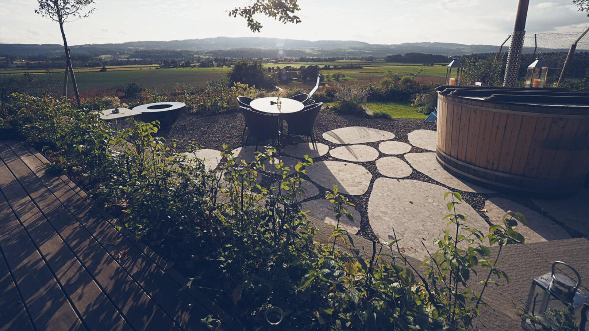 Terrasse mit modernen runden Möbeln, einem runden Holz-Whirlpool und Steinpflaster mit Blick auf eine malerische Landschaft mit Feldern und fernen Hügeln unter einem hellen Himmel.