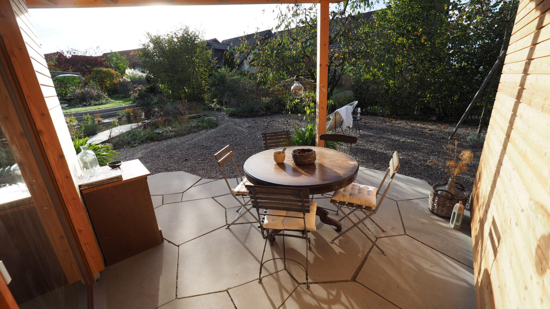 Ein runder Holztisch mit vier Stühlen steht auf einer Terrasse mit großen Steinfliesen und blickt auf einen Garten mit Bäumen und Sträuchern. Das Sonnenlicht wirft Schatten auf den Sitzbereich im Freien.