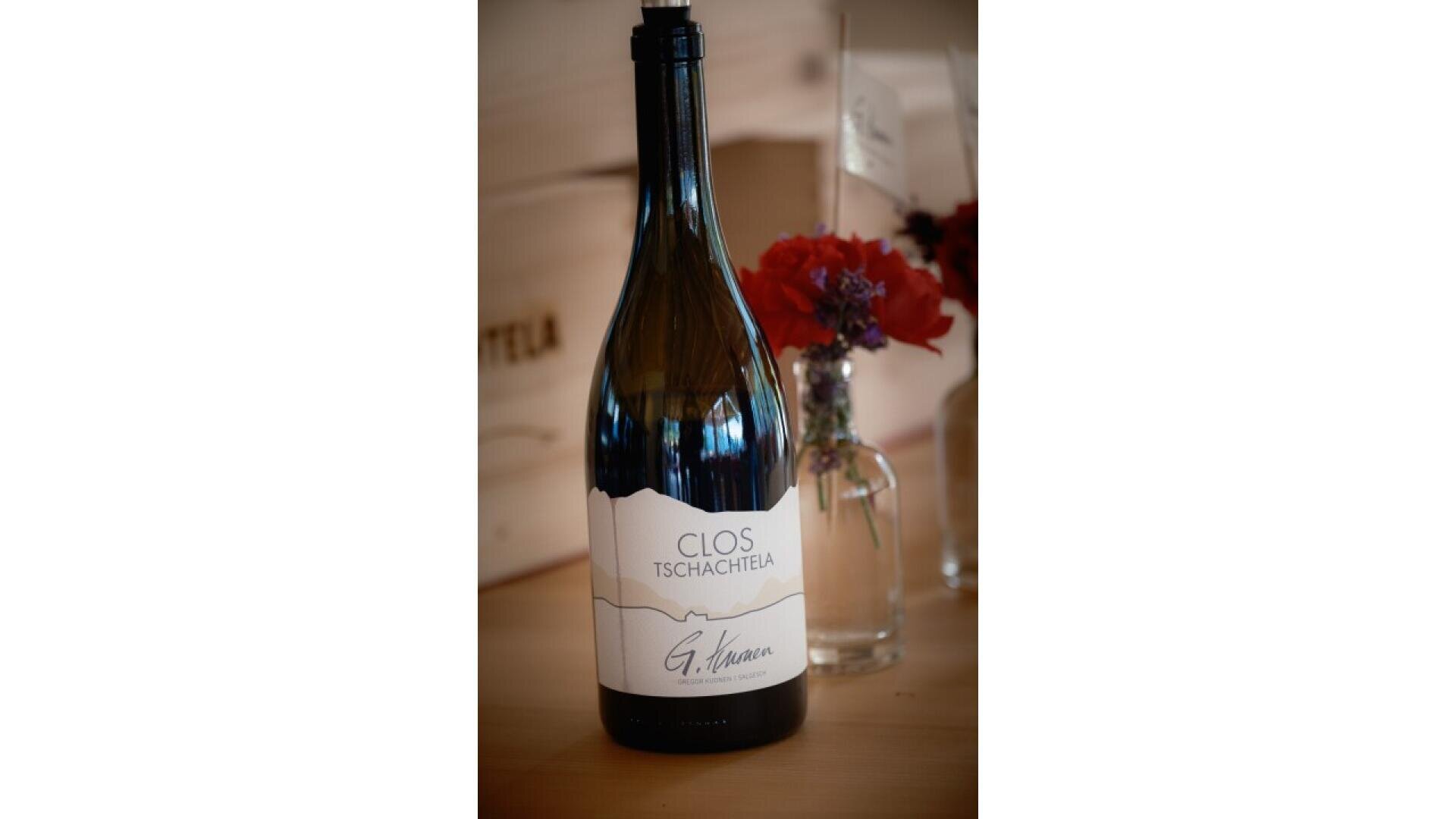 A bottle of Clos Tschachtela wine stands on a wooden surface next to a small glass vase containing red and purple flowers. The wine label features a mountain outline and the name G. Varone.