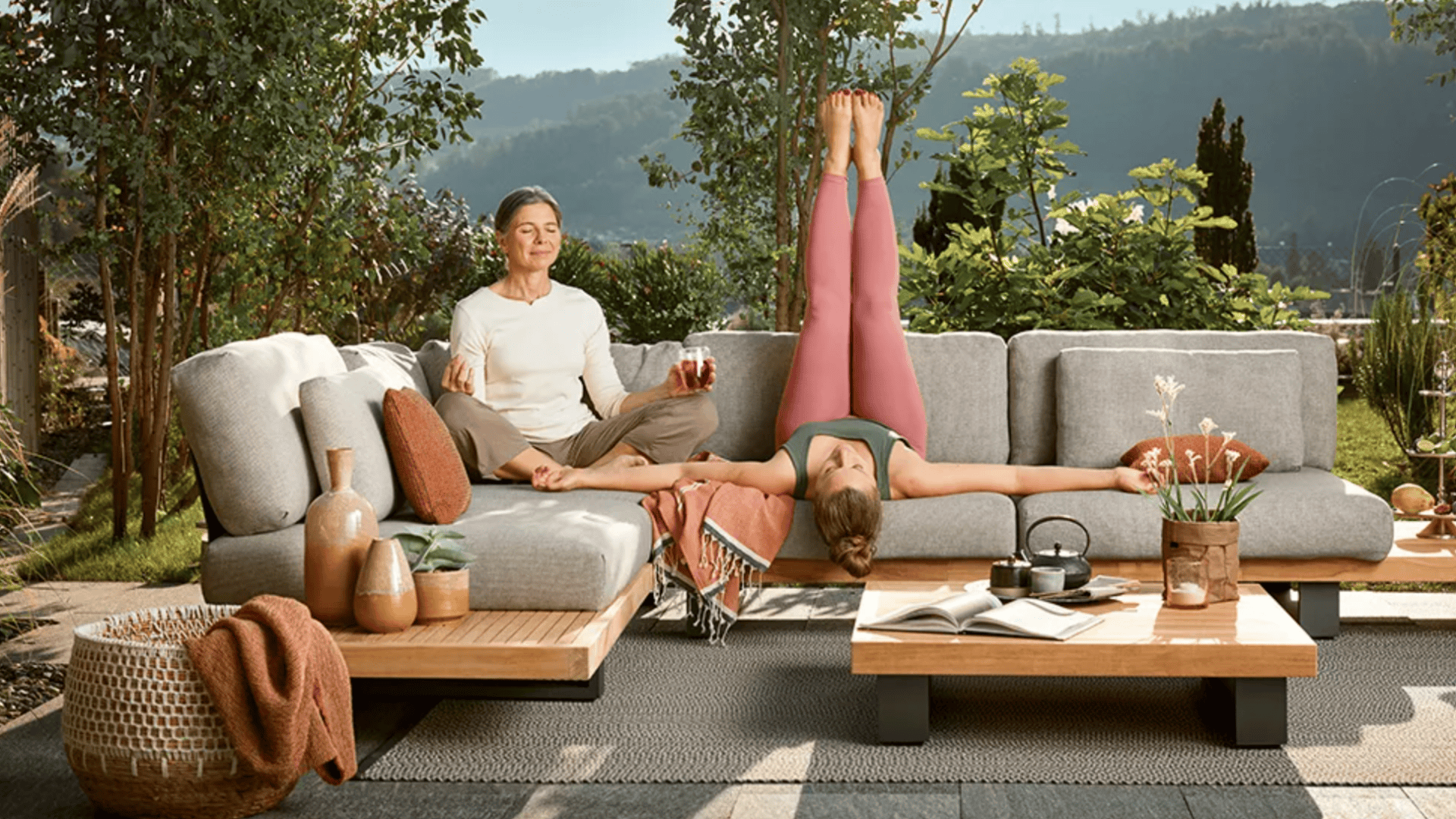 Zwei Personen entspannen sich auf einem Sofa im Freien; die eine sitzt im Schneidersitz und meditiert mit einem Getränk in der Hand, während die andere auf dem Rücken liegt und die Beine auf die Couch legt. Die Terrasse ist von Grünpflanzen und Dekorationsgegenständen umgeben.