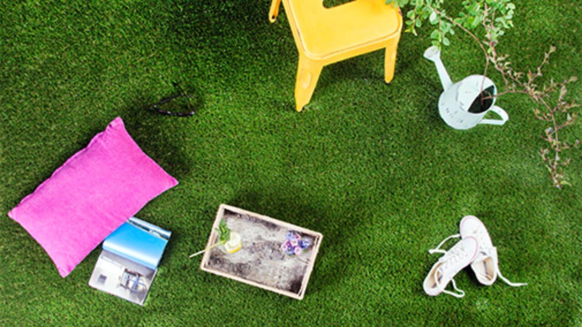A top-down view of a grassy area with a yellow chair, a pink pillow, a magazine, sunglasses, a wooden tray with a drink and flowers, white sneakers, and a watering can near a leafy plant.