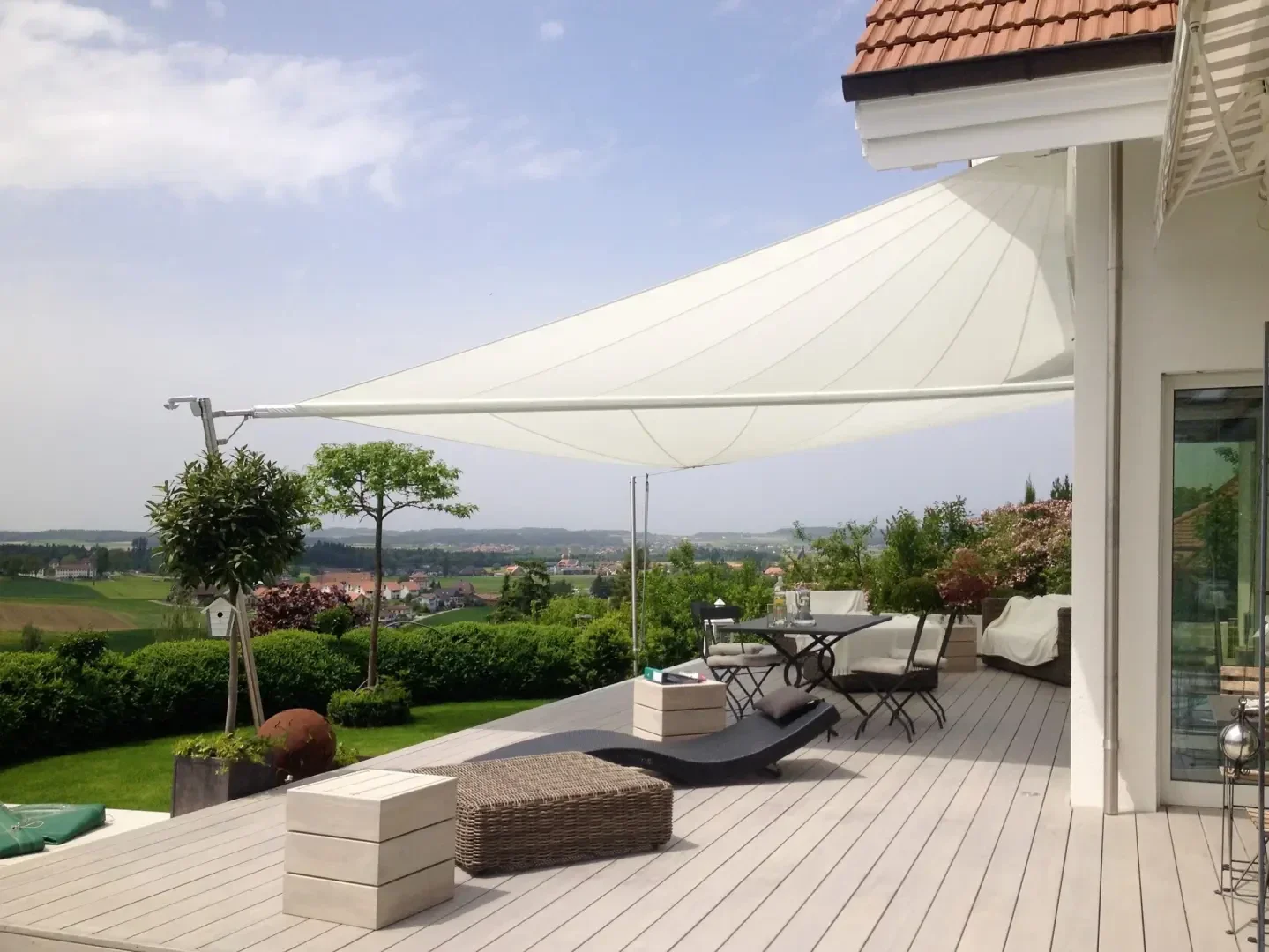 Modern patio with light wooden decking, stylish outdoor furniture, and a large white sunshade canopy. The area overlooks a scenic landscape with fields and distant hills under a partly cloudy sky.