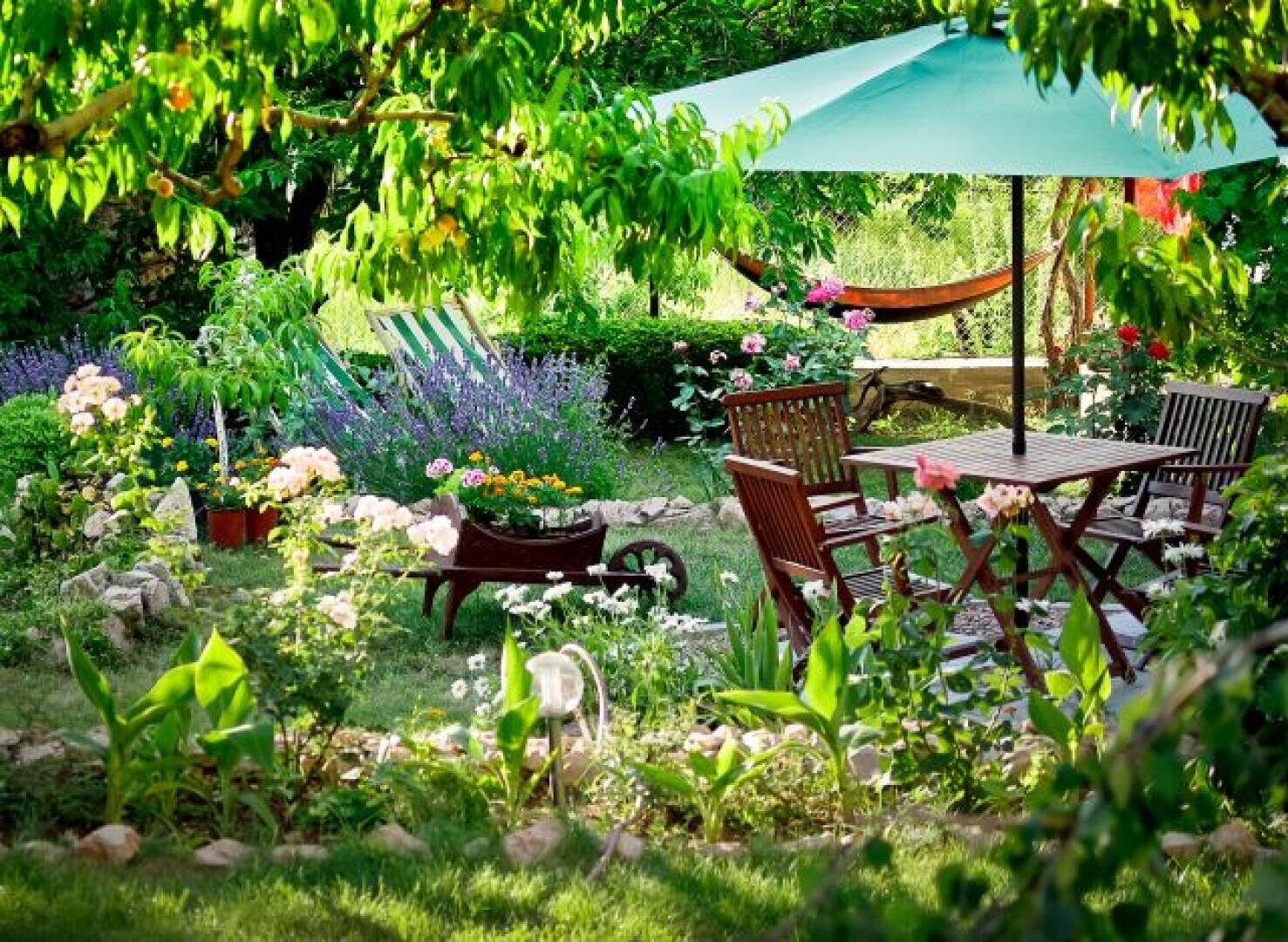 Ein üppiger Garten mit blühenden Blumen, grünen Pflanzen und Bäumen. Es gibt einen Holztisch mit Stühlen unter einem großen grünen Sonnenschirm, eine Hängematte im Hintergrund und eine mit Blumen gefüllte Schubkarre. Das Sonnenlicht fällt durch die Blätter.