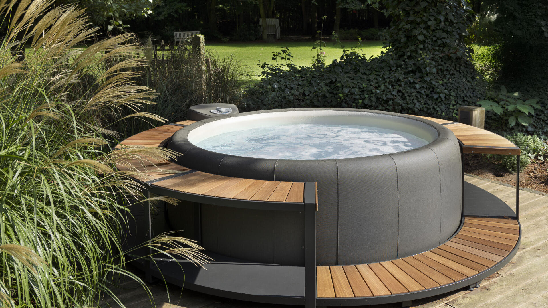 A round outdoor whirlpool with bubbling water, surrounded by a wooden deck, is located on a terrace next to tall grasses and lush greenery in a garden.