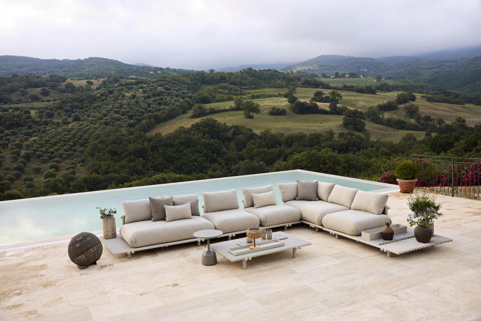 A modern outdoor seating area with beige sofas and a low table is located on a stone terrace next to the infinity pool with a view of rolling green hills under a cloudy sky.