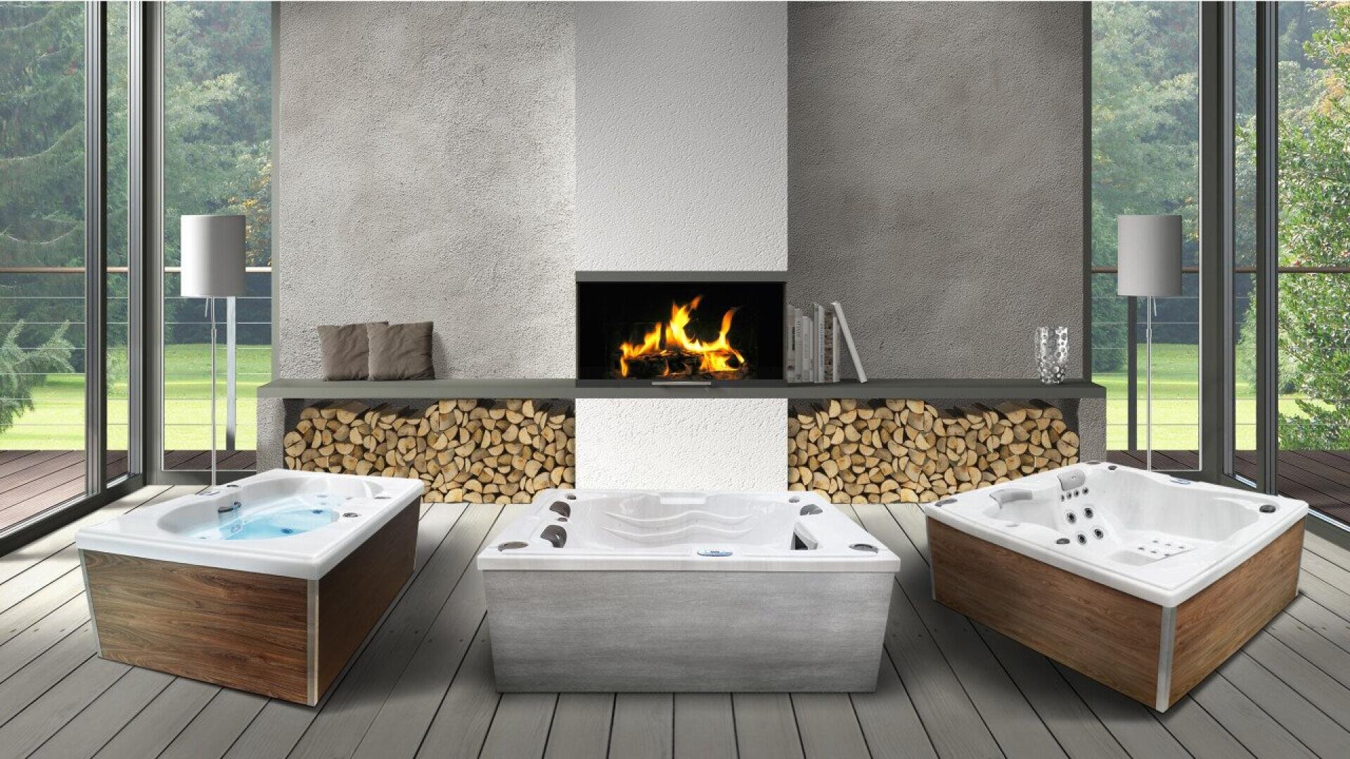 Three corner hot tubs are placed in a modern, glass-walled room with a central fireplace, stacked firewood, and a scenic view of trees outside.