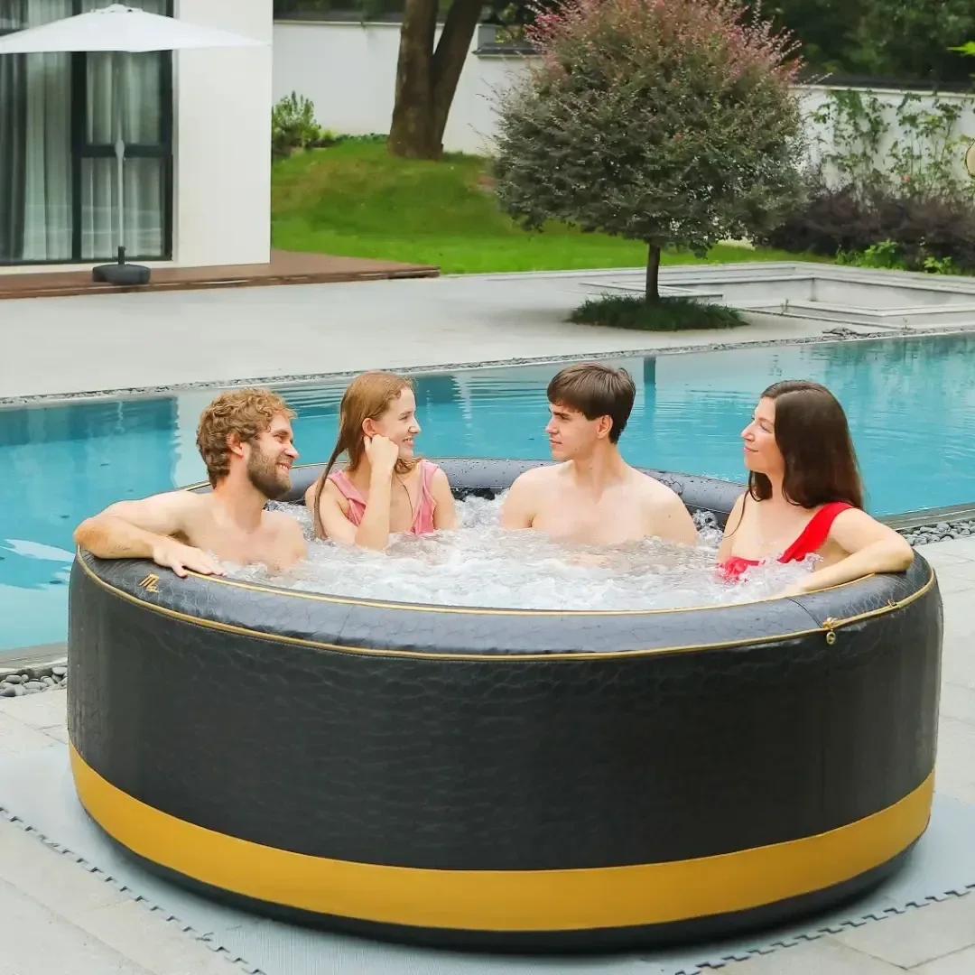 Vier Personen sitzen und lächeln in einem runden, schwarz-goldenen aufblasbaren Whirlpool neben einem Swimmingpool in einem Hinterhof, mit Grünpflanzen und einem modernen weißen Gebäude im Hintergrund.