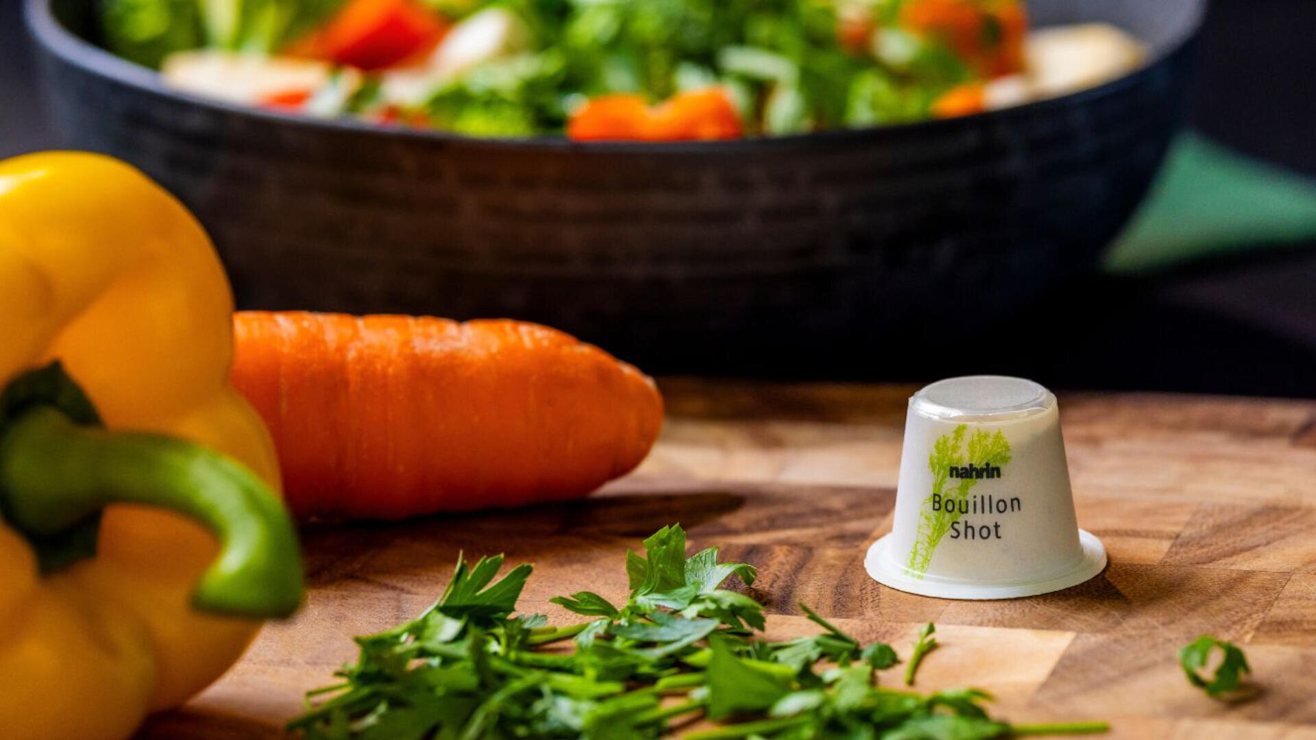 Eine gelbe Paprika, eine Karotte und gehackte Petersilie liegen auf einem hölzernen Schneidebrett. In der Nähe steht ein kleiner Behälter mit der Aufschrift "Bouillon Shot", im Hintergrund eine Schüssel mit gemischtem Gemüse.
