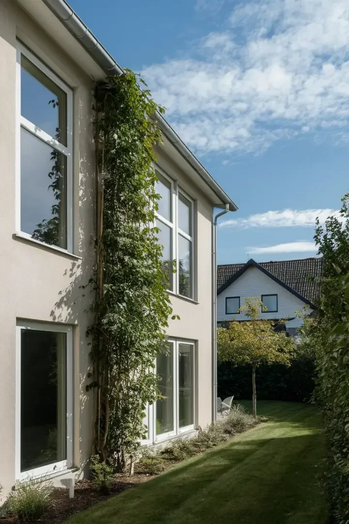 Ein modernes zweistöckiges Haus mit großen Fenstern und einer vertikalen grünen Ranke, die an der Wand entlangwächst, umrahmt von einem ordentlich getrimmten Rasen und einem kleinen Garten, unter einem teilweise bewölkten blauen Himmel. Im Hintergrund ist ein weiteres Haus zu sehen.