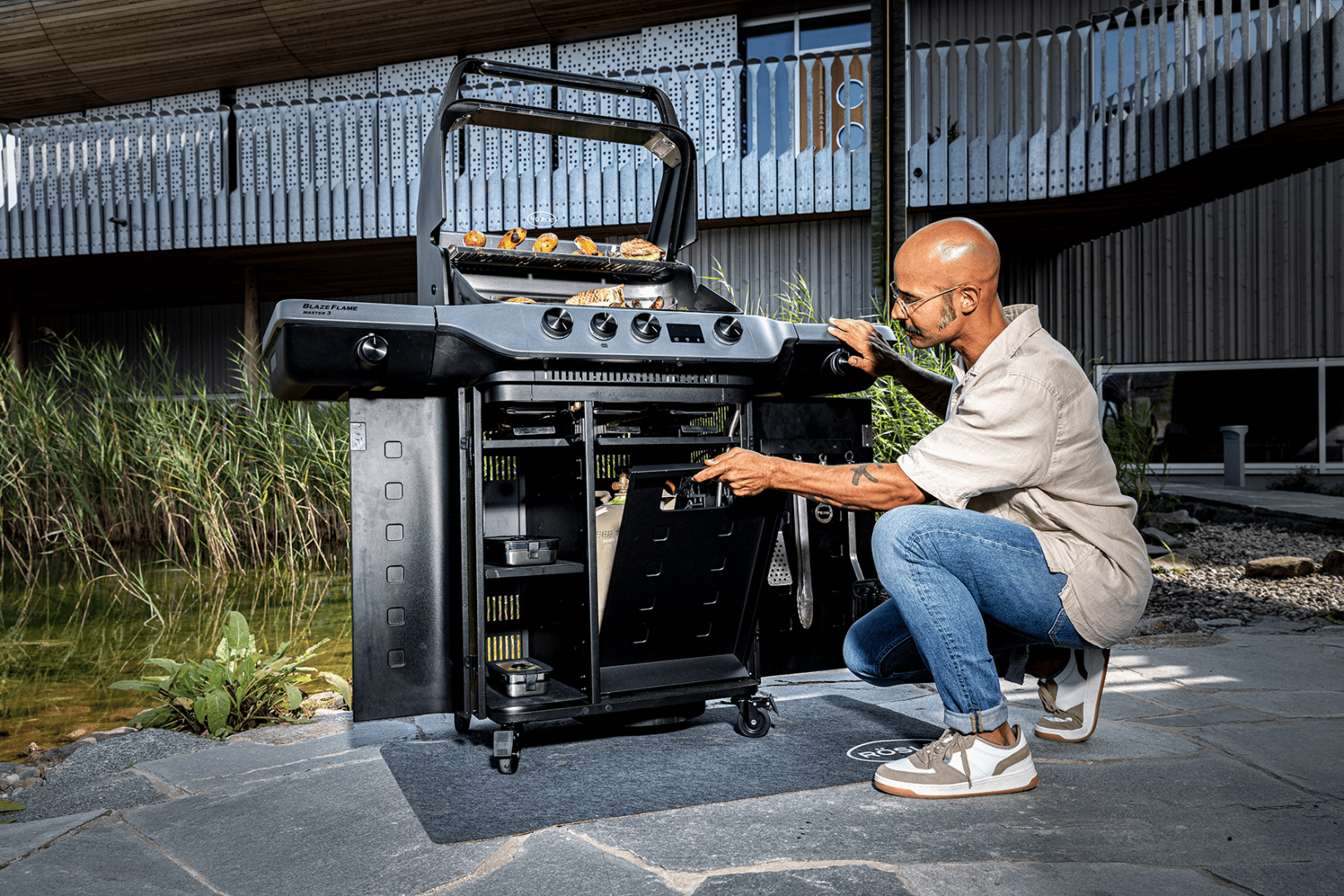 Eine Person kniet neben einem schwarzen Außengrill und stellt ein Bauteil unter dem Grill ein. Das Essen wird auf dem Grill gegart. Die Szene spielt auf einer Steinterrasse neben einem Teich und einem modernen Gebäude.