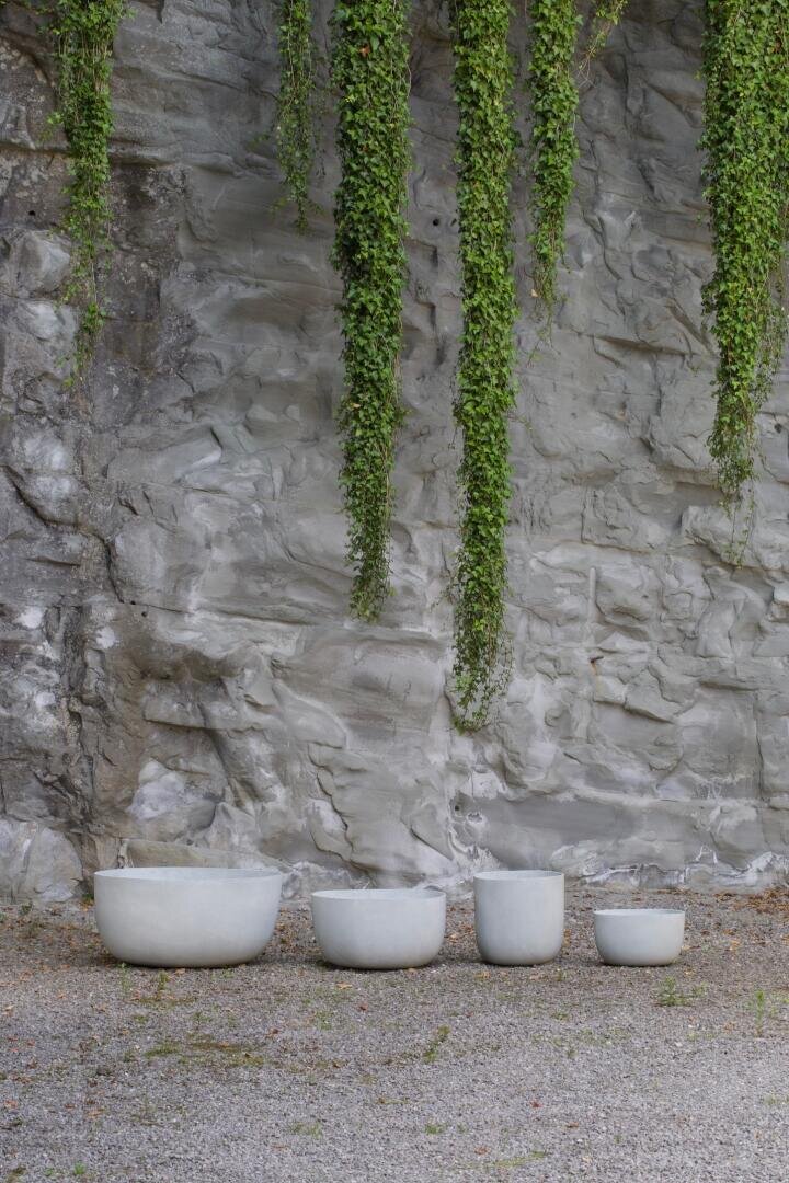 Four white, minimalist ceramic pots of different sizes stand in a row on gravel in front of a gray stone wall, from which long green vines hang down.