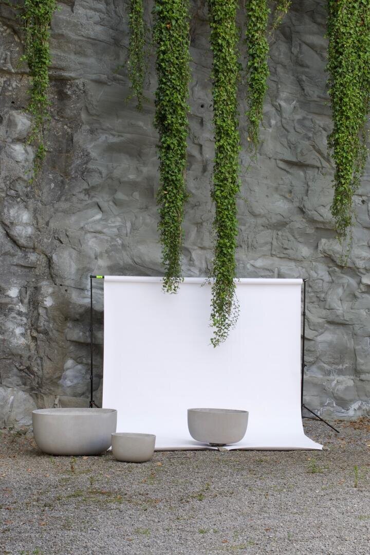 A white backdrop stands against a rocky wall with trailing green vines above, while three large, empty gray plant pots are placed on gravel in front of the backdrop.