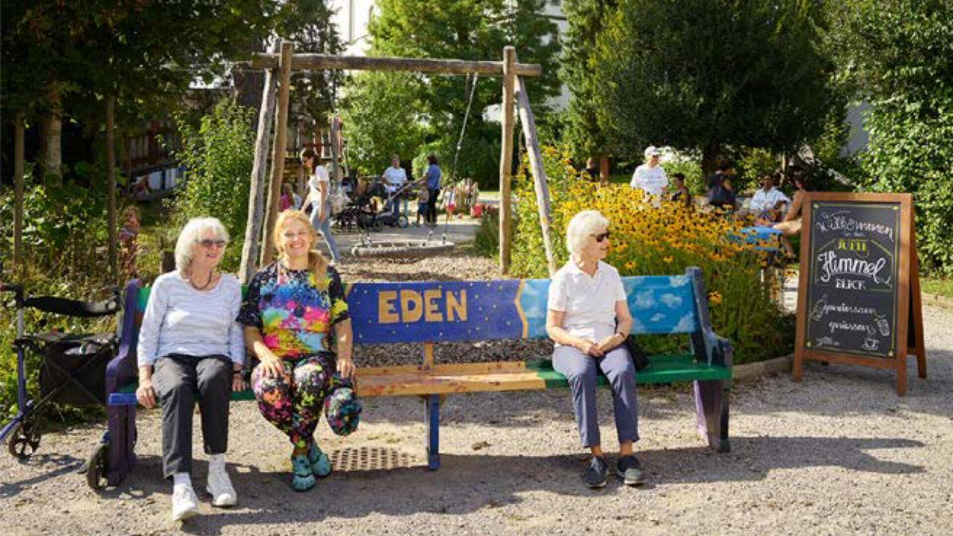 Drei Frauen sitzen auf einer bunten Bank mit der Aufschrift EDEN in einem sonnigen Park. Hinter ihnen versammeln sich Menschen in der Nähe von Schaukeln und Blumen, und auf der rechten Seite ist ein Kreideschild zu sehen.