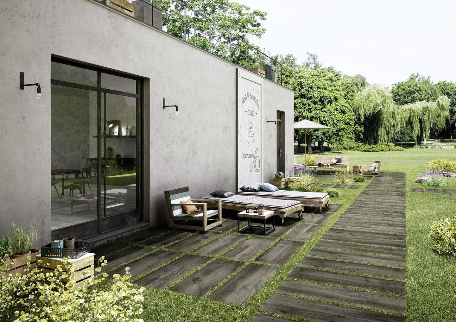 Modern patio with wooden outdoor furniture and lounge chairs on a tiled pathway, next to a building with large glass doors, overlooking a lush green garden with trees and flowers.