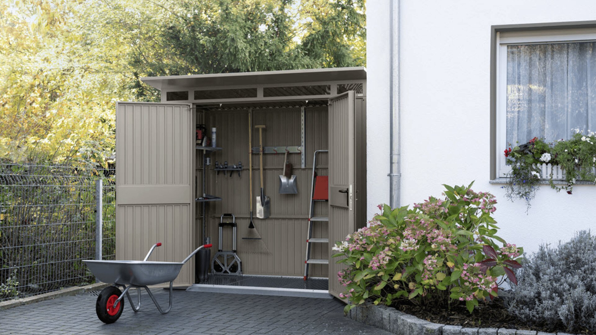 Ein kleines Metall-Gartenhaus mit offenen Flügeltüren steht neben einem weißen Haus. Im Inneren befinden sich hängende Werkzeuge und eine Leiter. Draußen steht eine Schubkarre, und in der Nähe befinden sich Blumen, Sträucher und ein Fenster mit einem Blumenkasten.