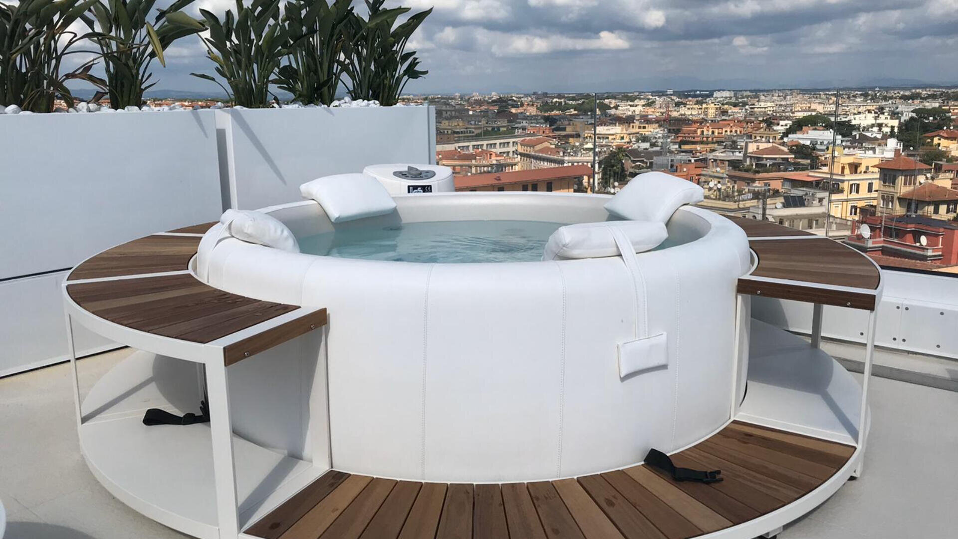 Ein moderner weißer, runder Whirlpool mit hölzernen Akzenten und eingebauten Bänken steht auf einem Dach, umgeben von Pflanzen und mit Blick auf eine Stadtlandschaft unter einem teilweise bewölkten Himmel.