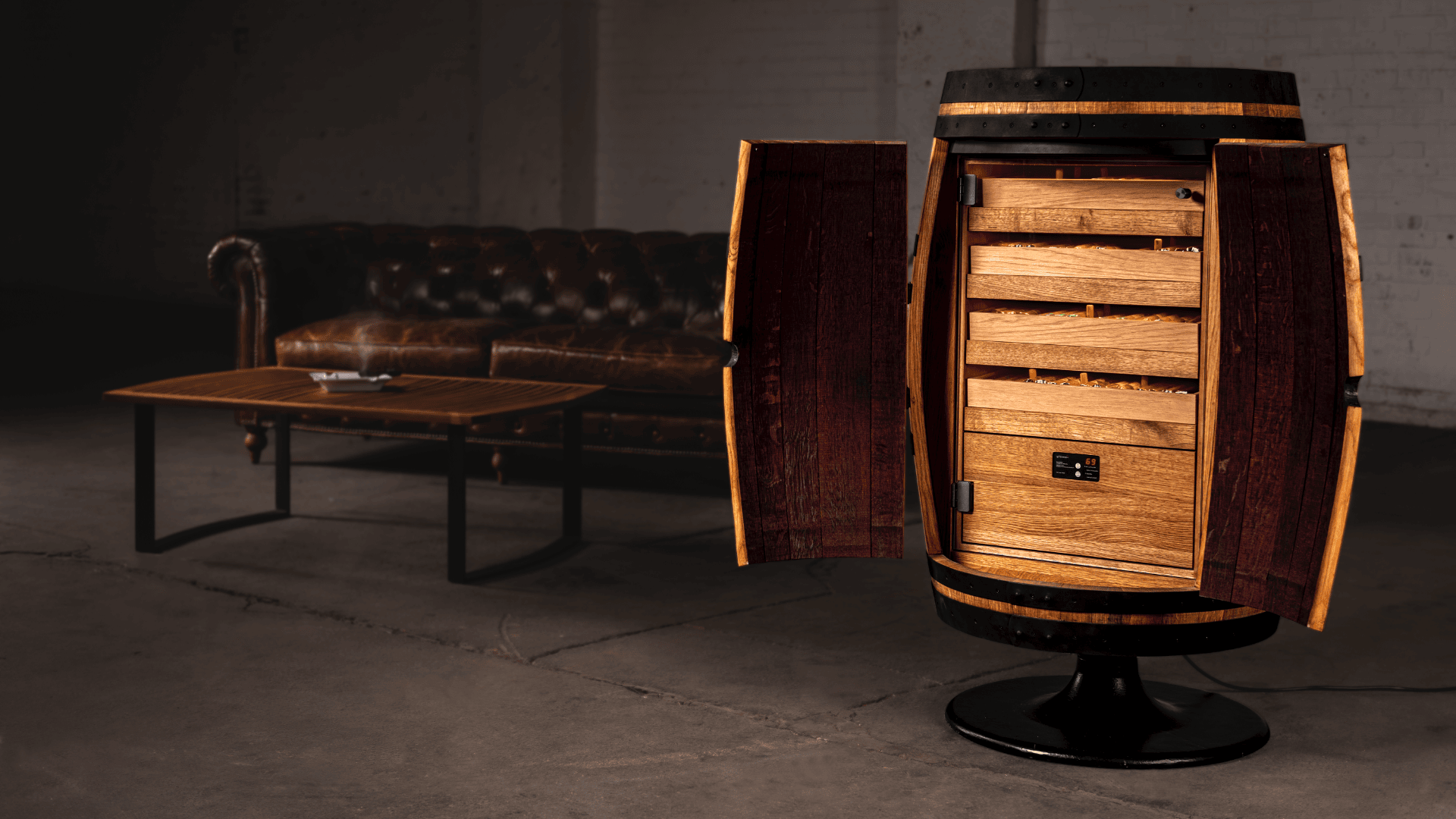 Ein als Humidor umfunktioniertes Holzfass mit offenen Türen, die den Blick auf Regale im Inneren freigeben, steht auf einem schwarzen Sockel in einem Loft mit einer Ledercouch und einem hölzernen Couchtisch im Hintergrund.