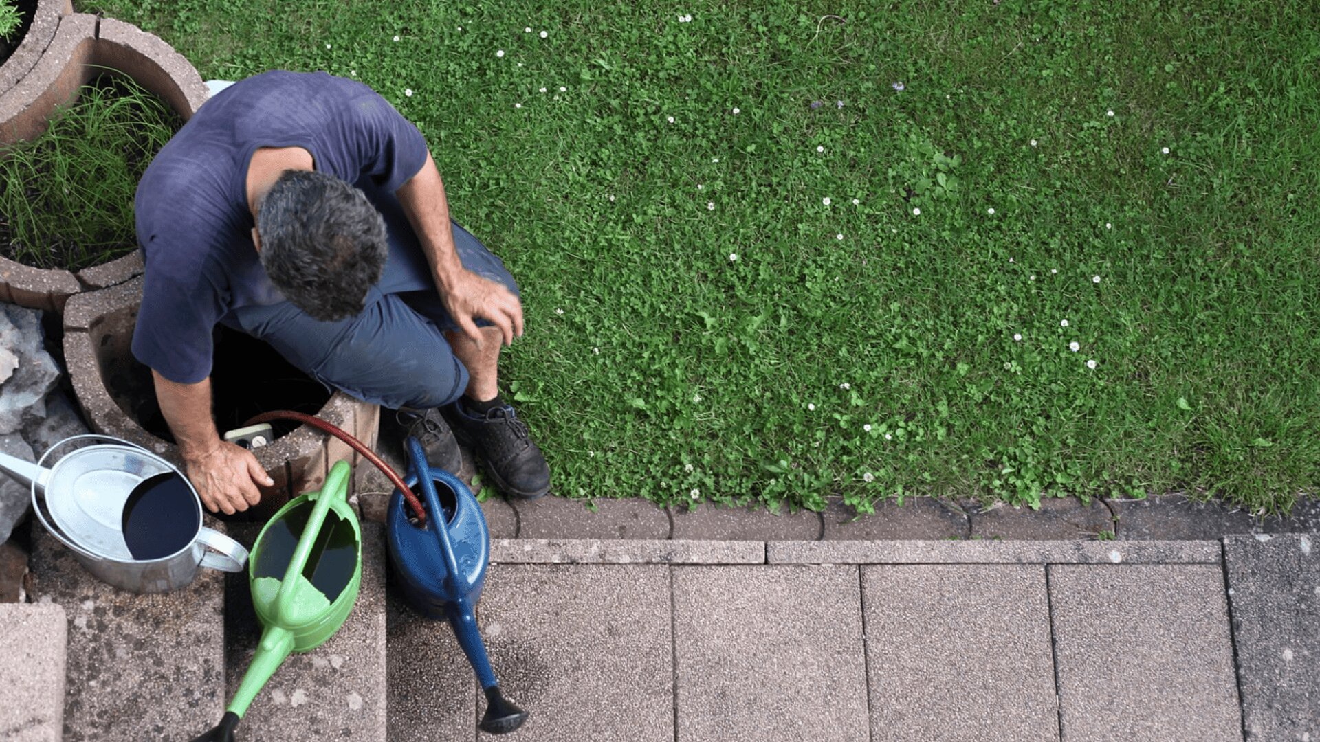 Eine Person mit dunklem Haar füllt blaue und grüne Gießkannen aus einem Eimer in der Nähe eines grasbewachsenen Rasens, der auf Steinfliesen neben einem Stück grünen Grases steht. Die Szene ist von oben betrachtet.