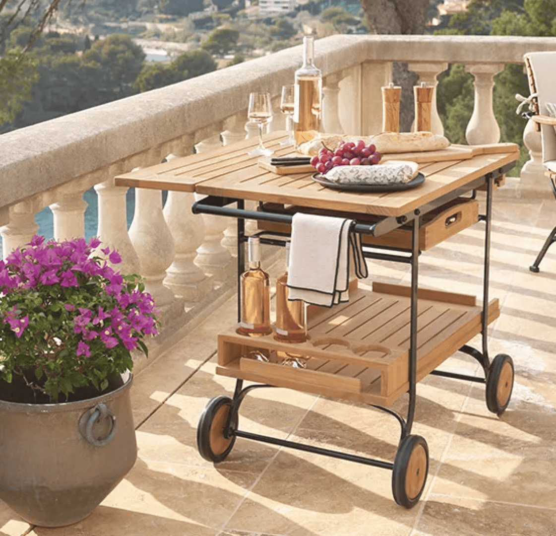 A wooden serving cart with wine, glasses, bread, and grapes sits on a sunny outdoor terrace with a scenic view. A potted plant with purple flowers is nearby, and a white towel hangs from the cart.